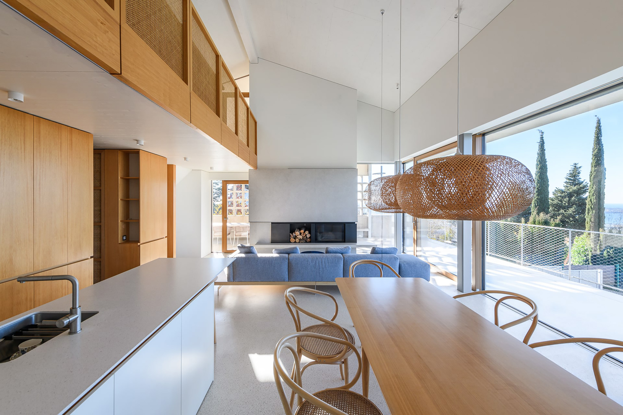 Open kitchen and dining area with oak island, cane pendant lamps and a blue sofa facing a fireplace, cypress trees outside