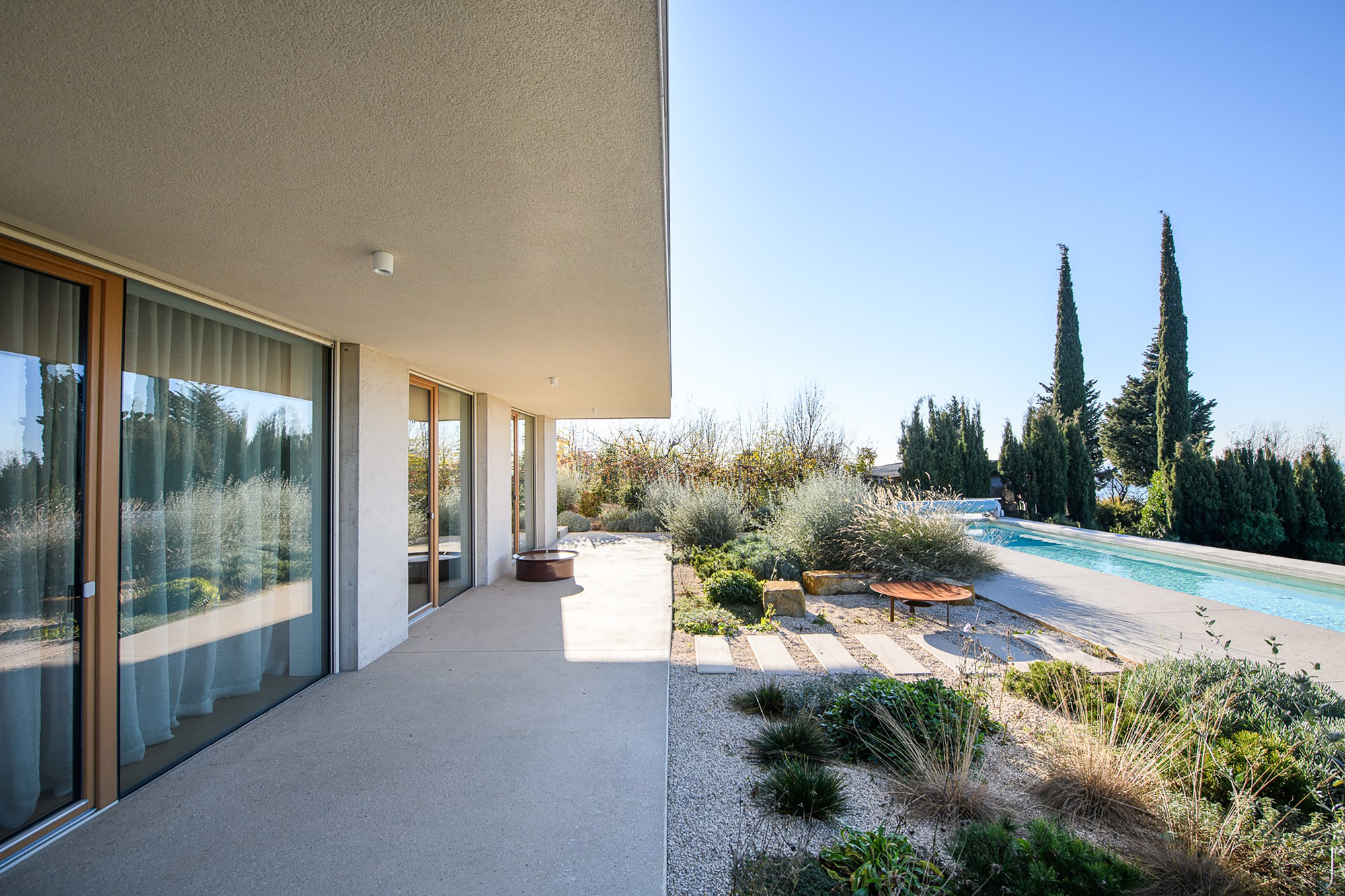 Covered concrete terrace with timber-framed glass doors and sheer curtains, overlooking a gravel garden of grasses and a lap pool in midday sun