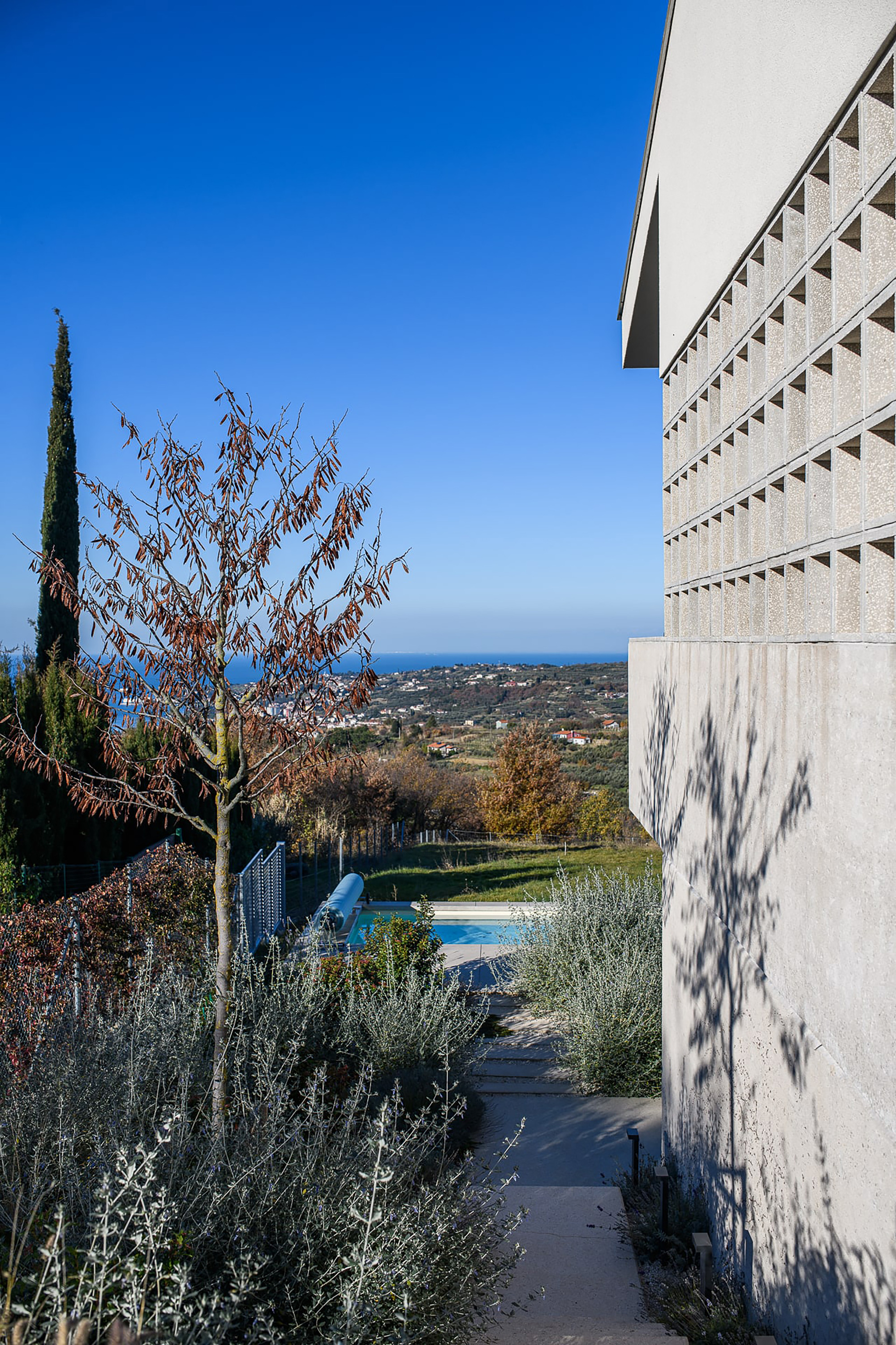 Side facade with perforated concrete block screen above a stepped garden path, with a bare tree casting shadows toward the sea