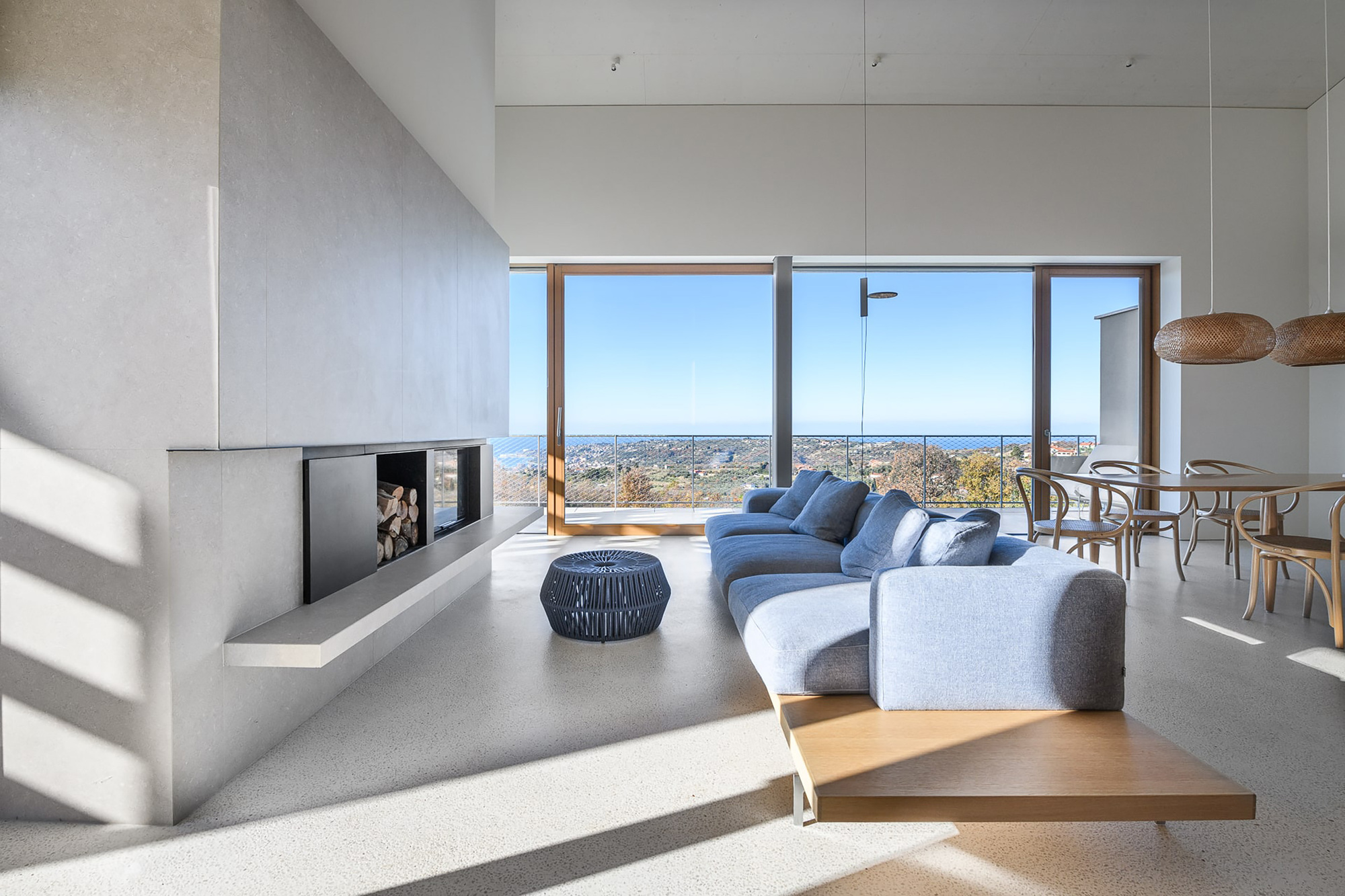 Living room with concrete fireplace, low blue sofa and floor-to-ceiling glazing framing a sea view, strong sunlight casting diagonal shadows across the floor