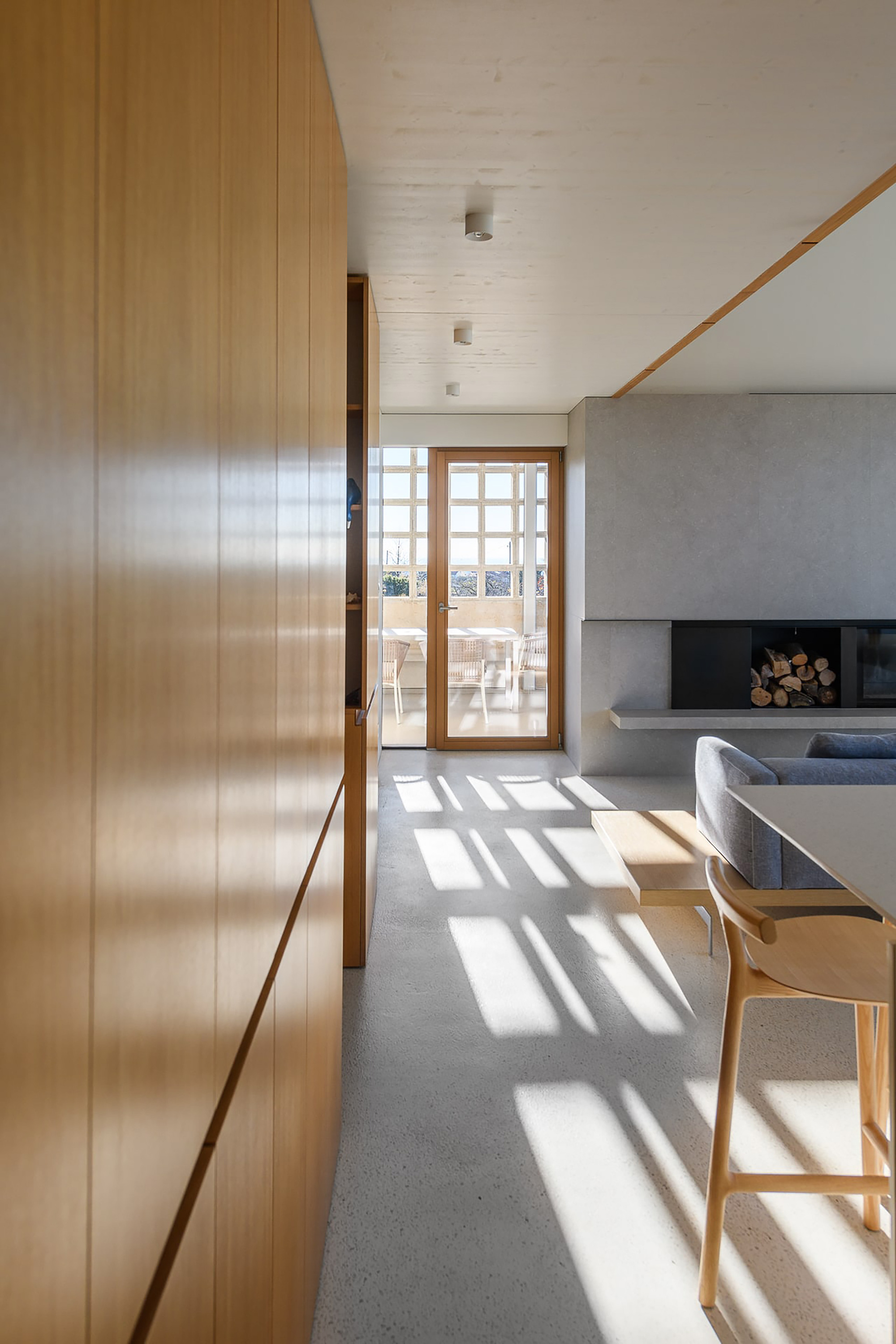 Oak cabinet wall running along a corridor toward a glazed timber door, with window shadows striping the terrazzo floor