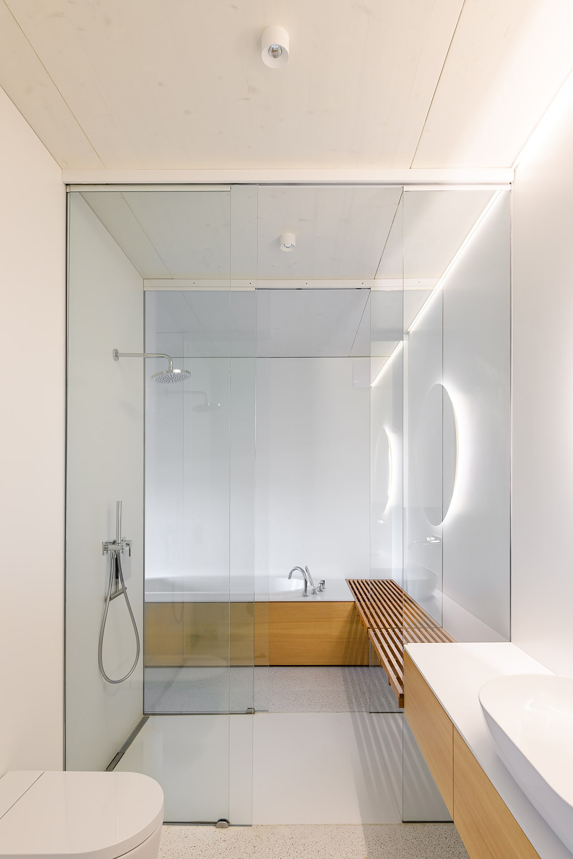 Bathroom with glass-enclosed shower, timber-clad bathtub, slatted oak bench and an oval backlit mirror beside a floating vanity