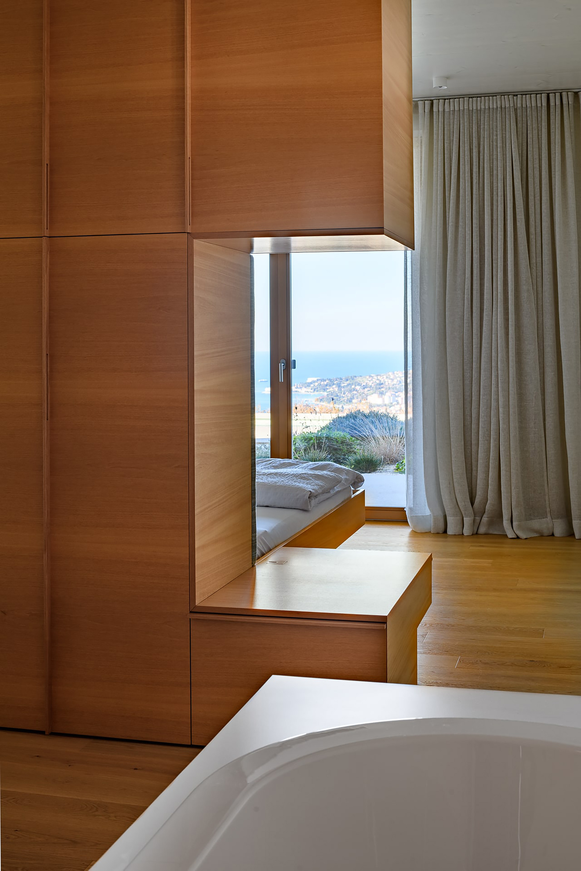 Oak-panelled bedroom nook with a window bench framing a sea view, a linen curtain and a bathtub edge in the foreground