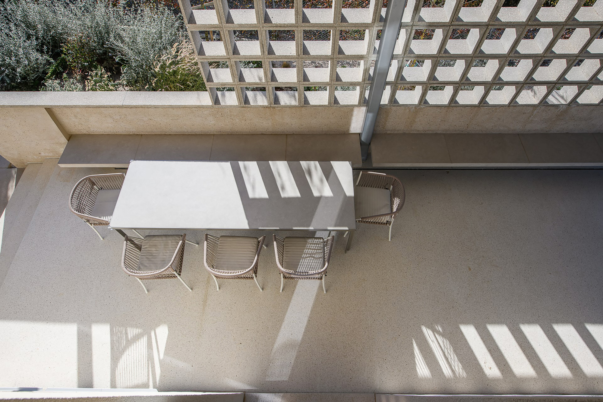 Top-down view of a terrace dining table and rope chairs beside a perforated concrete block screen casting striped afternoon shadows