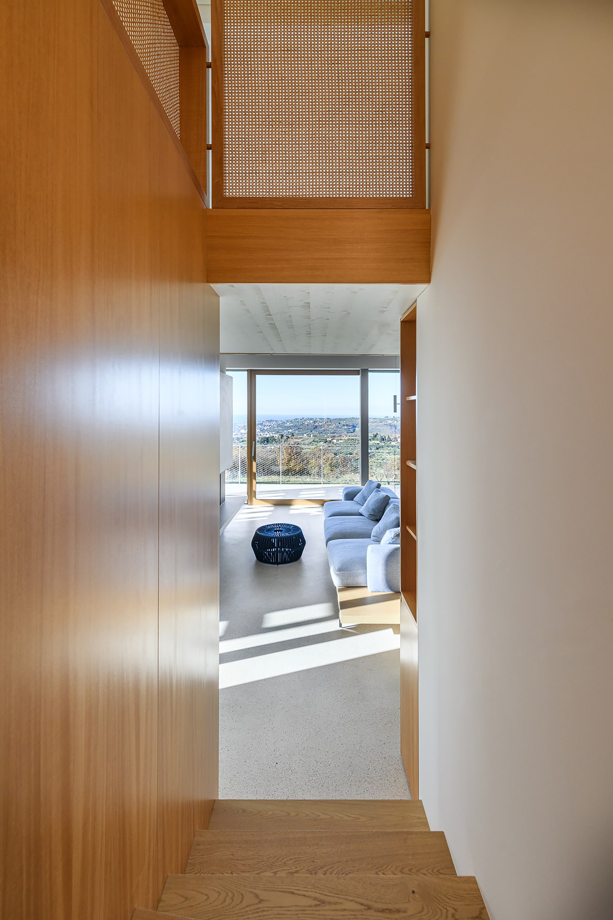 Oak-lined stairwell looking toward a living room with a blue sofa and hillside view, a cane-panelled balustrade overhead
