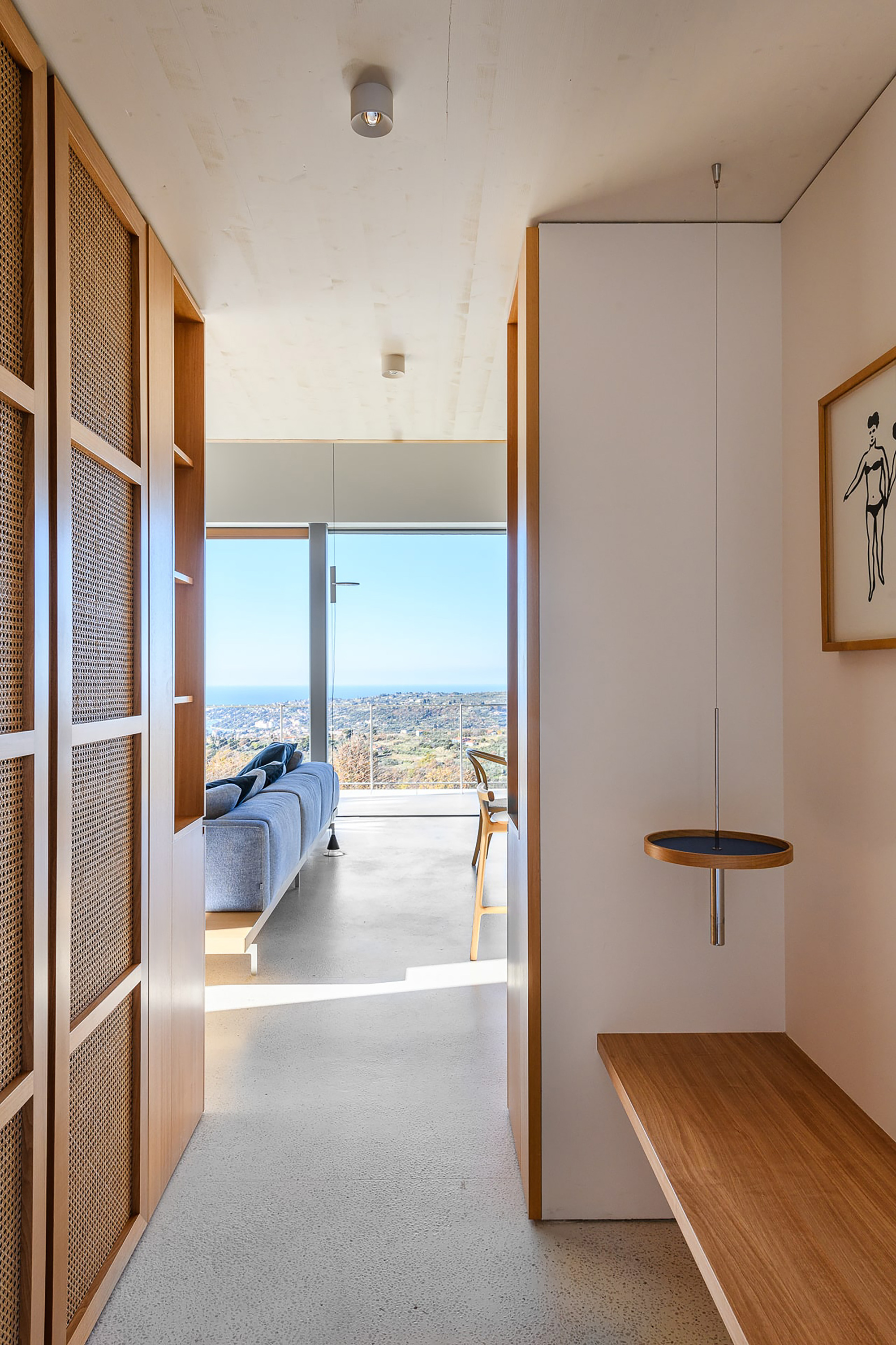 Entry passage with cane-panelled cabinet doors and an oak bench leading to a blue sofa and glazed sea view in bright sun