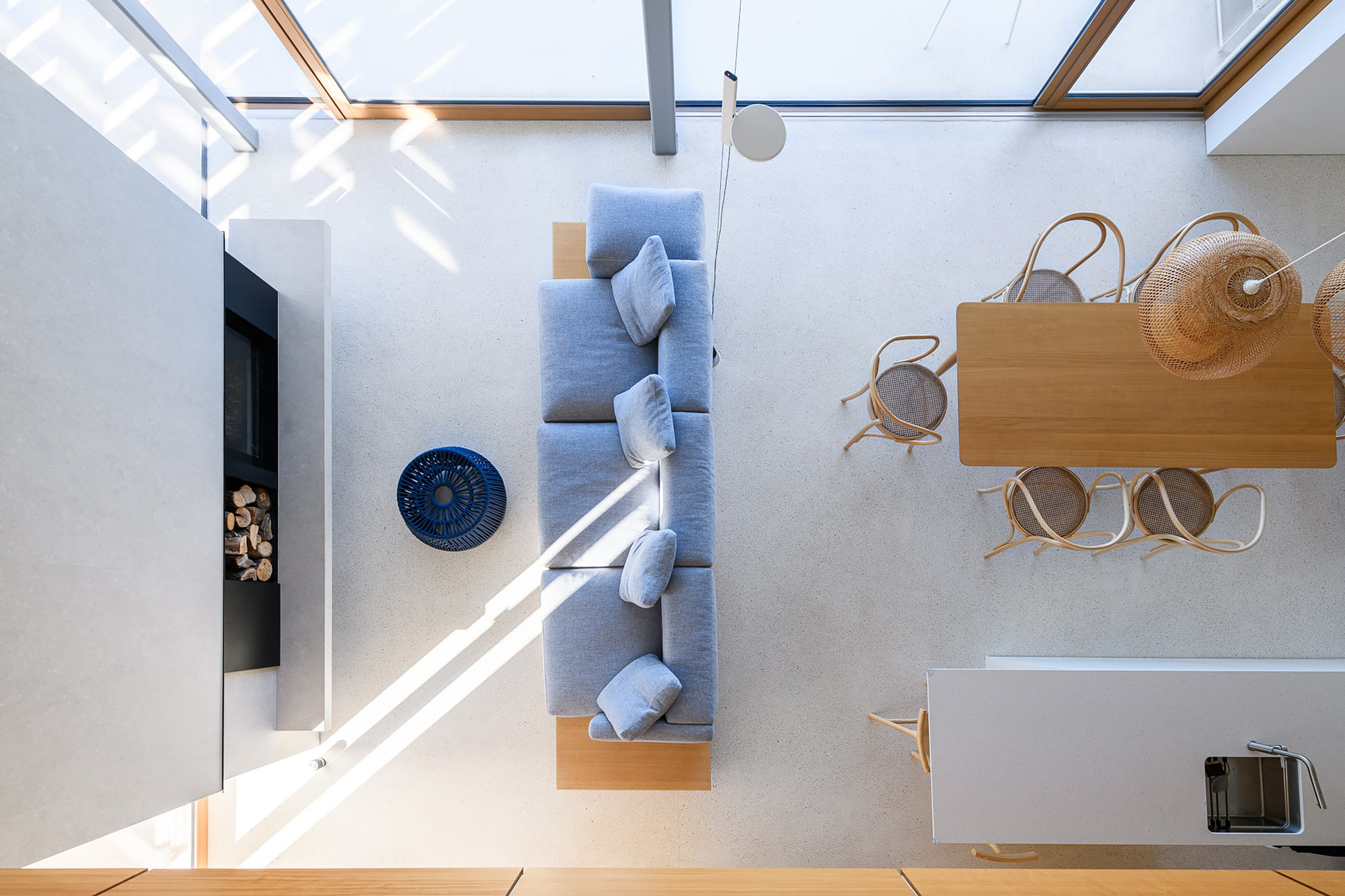 Top-down view of a blue-grey sofa, round blue pouf and oak dining table with bentwood chairs in streaks of sunlight