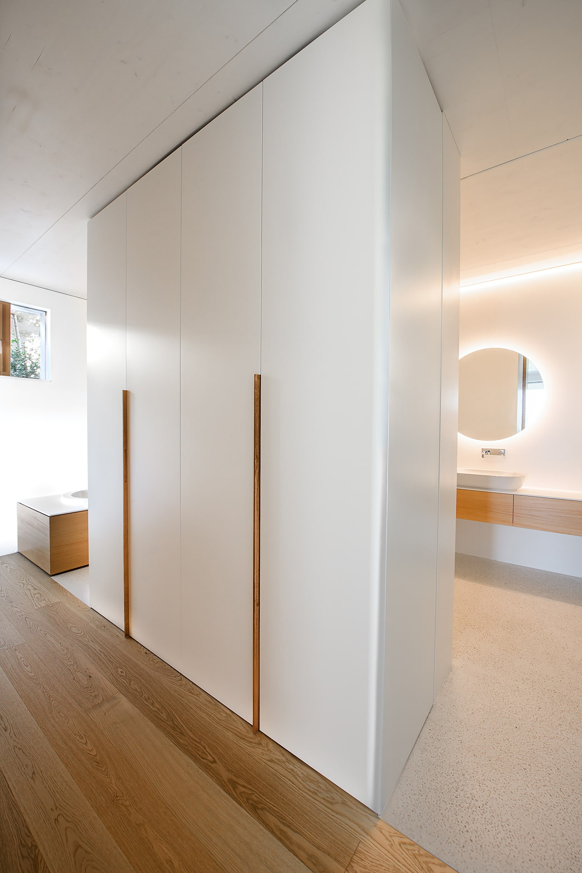 White built-in wardrobe with slim oak pull handles beside an oak floor, with a round backlit mirror in the bathroom beyond