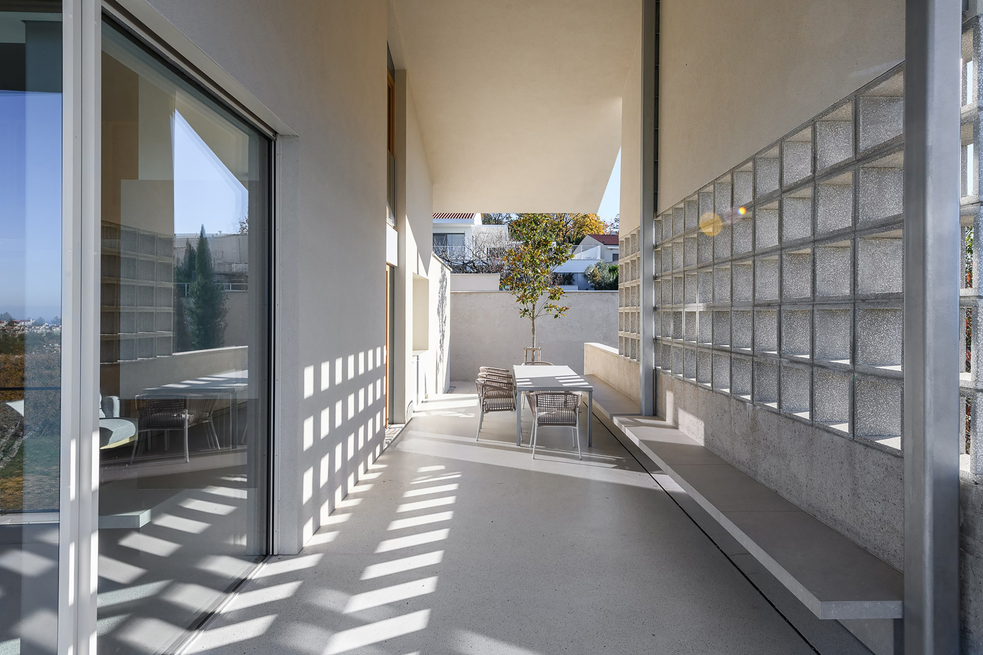 Covered terrace with a glass block wall, built-in concrete bench and dining table, dappled with sunlight shadows on the floor