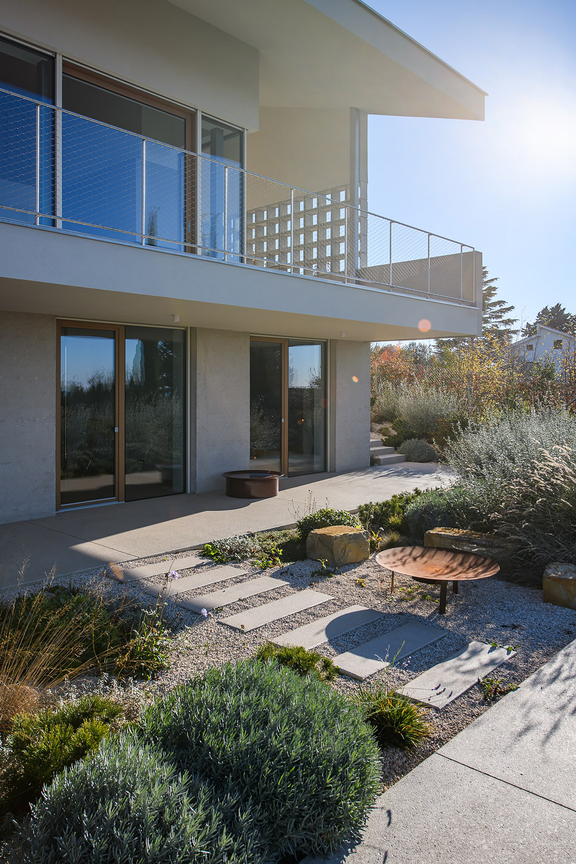 Garden facade with a balcony railing above sliding glass doors, stepping stones crossing a gravel bed of grasses and shrubs in low sun