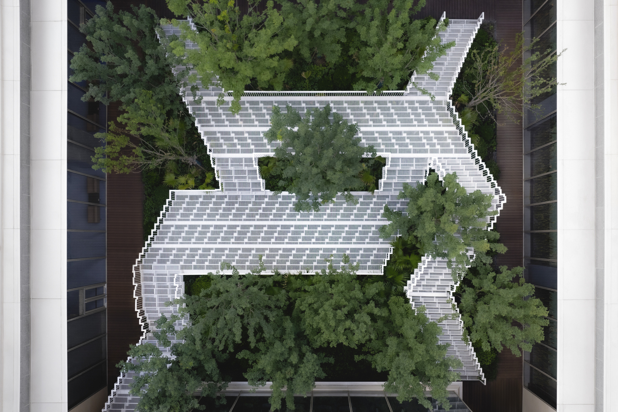 Top-down view of the zigzag white slatted walkway threading through a courtyard of dense green tree canopies between glazed facades