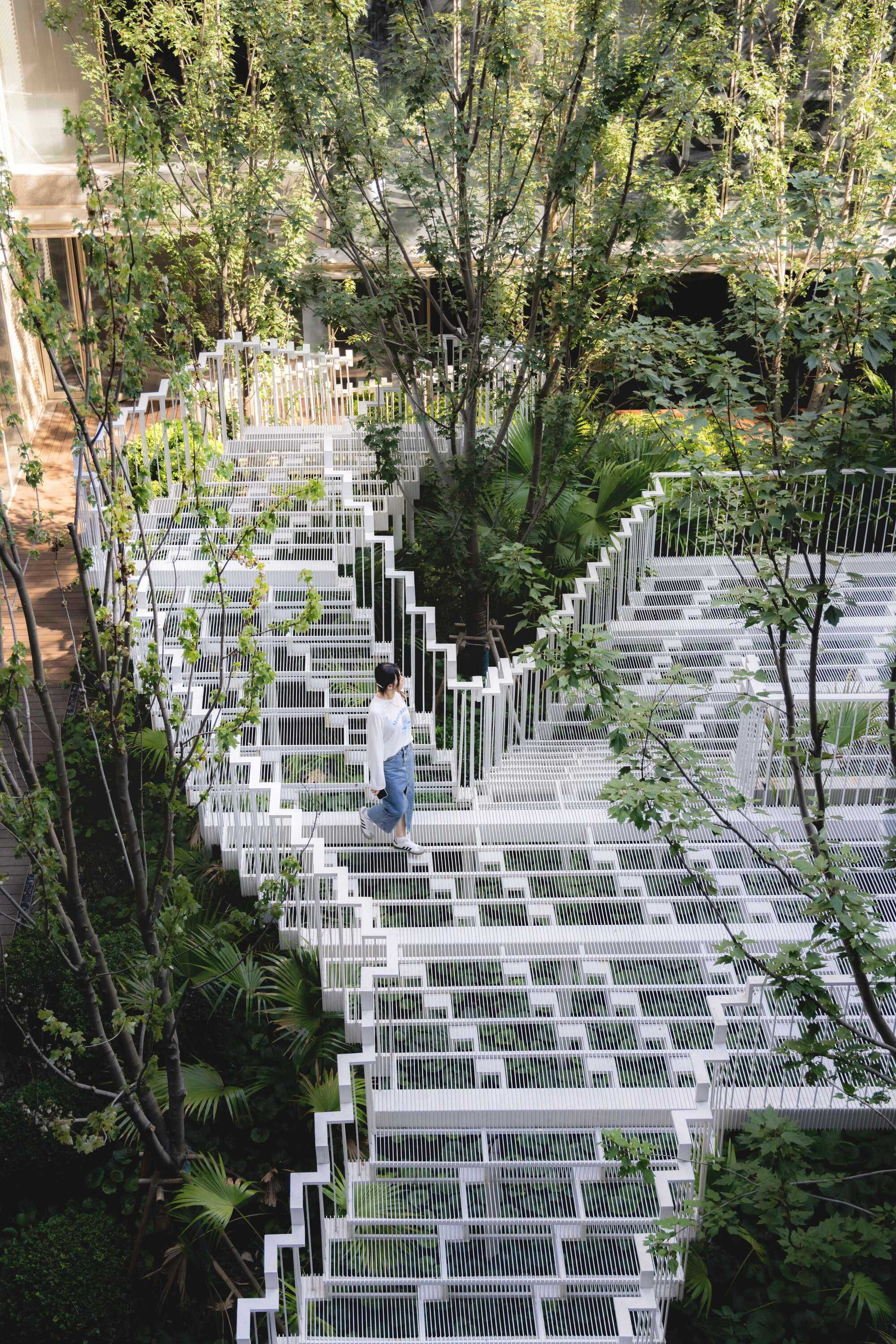 Woman walking down a stepped white grating walkway among tree trunks and palms in a courtyard