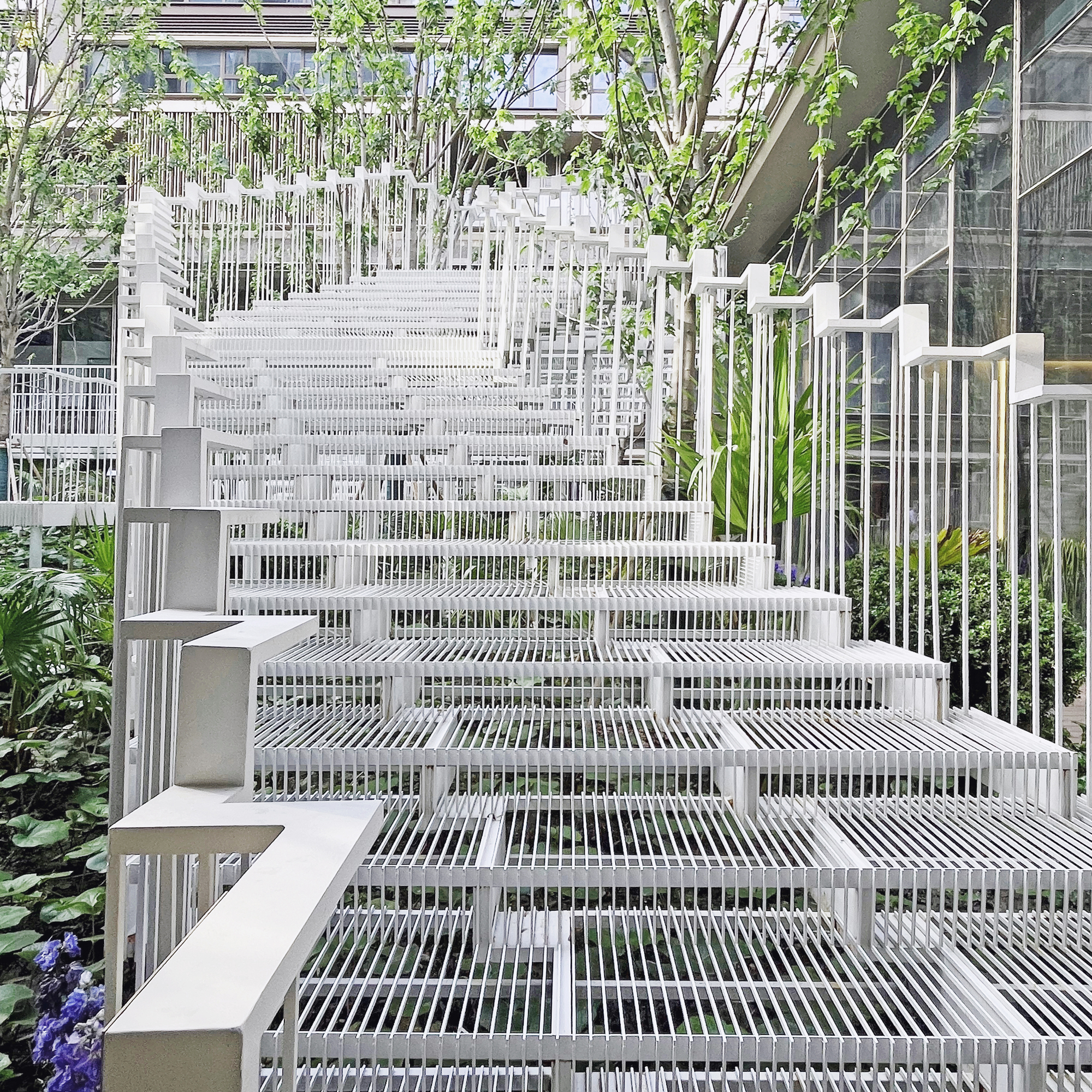 Frontal view of the white steel slatted stair rising between railings, with glass facade and young trees at right