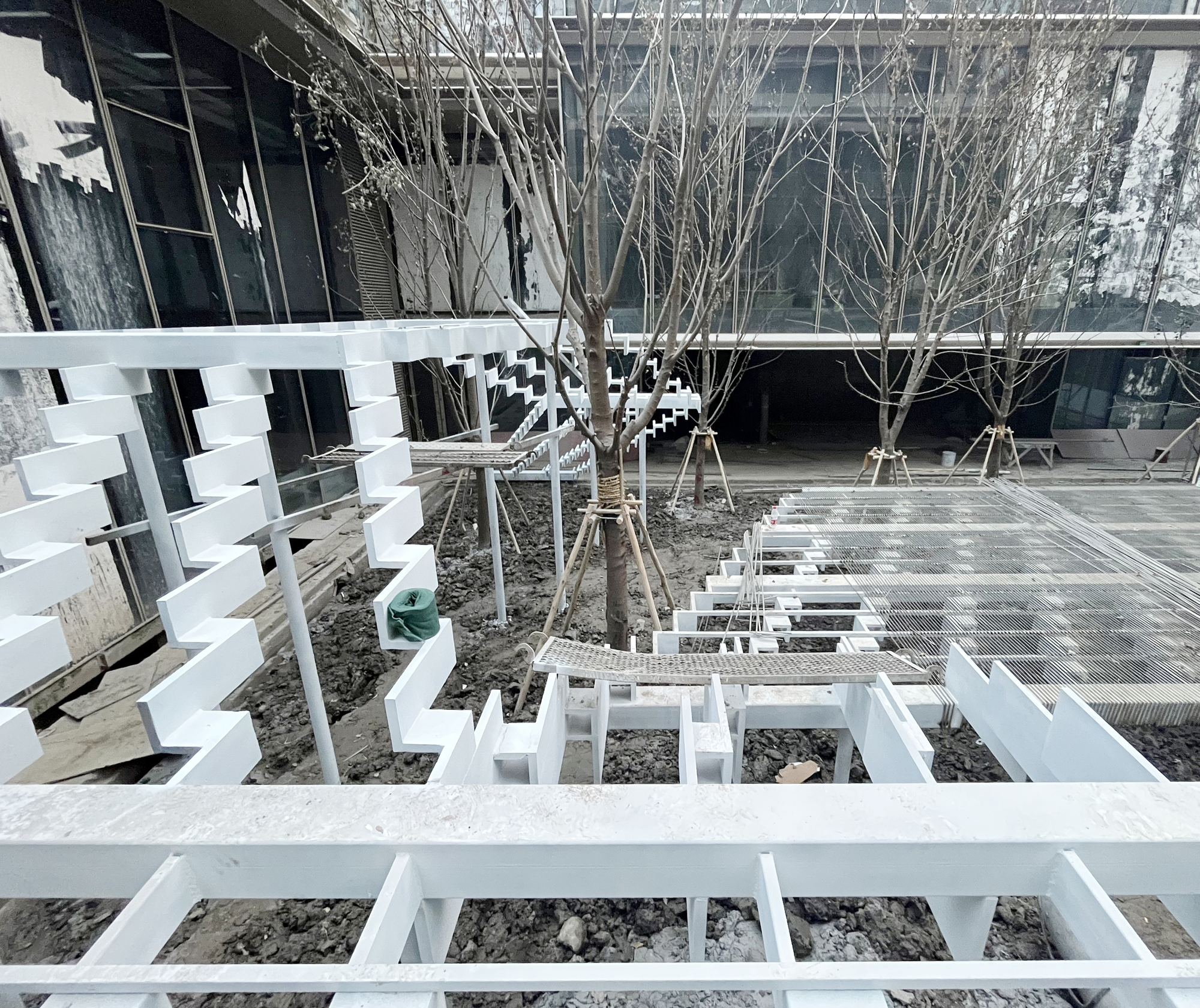 White stepped timber framework over bare soil in a courtyard of young leafless trees, with glass facades behind in winter