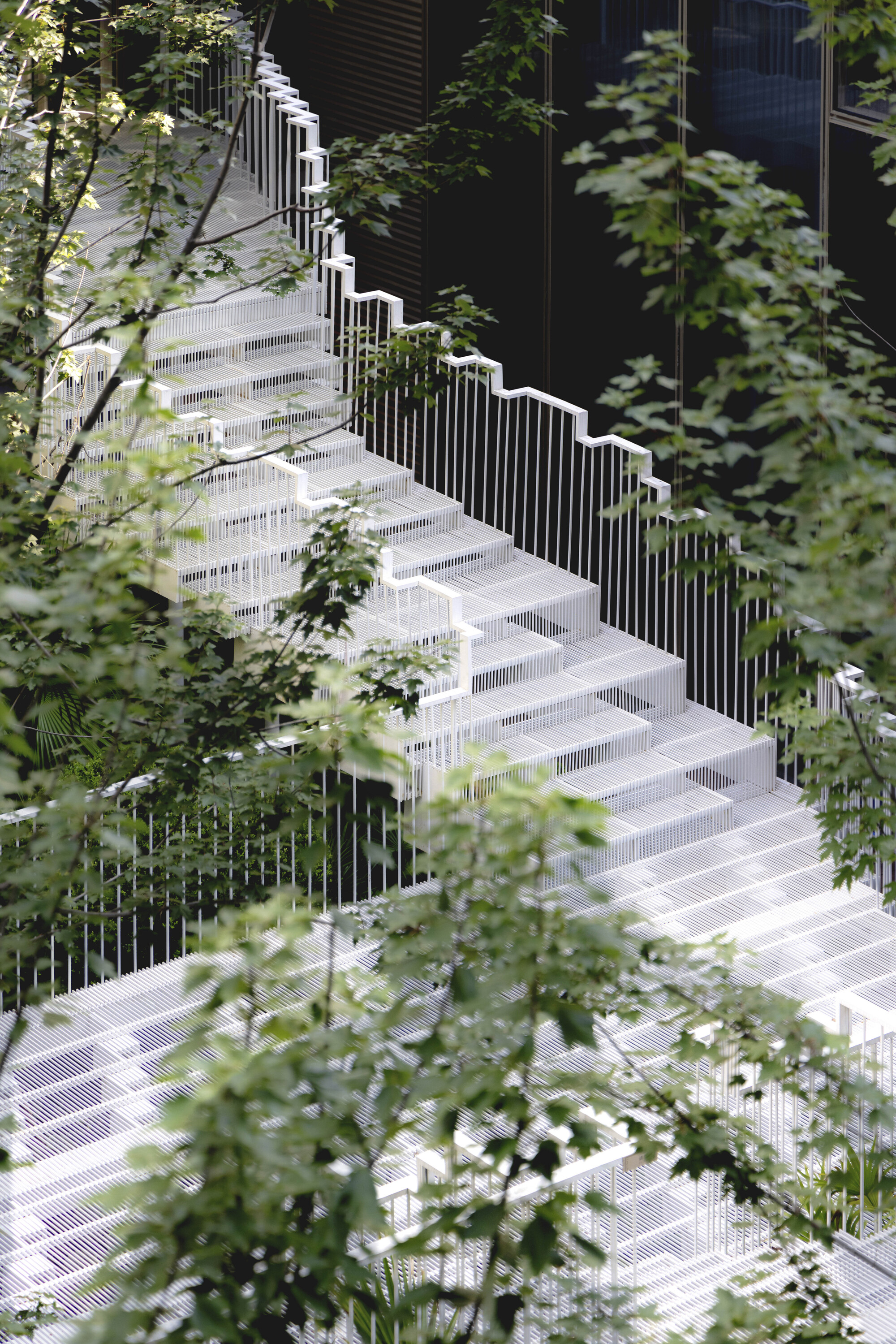 White perforated stairs with fine vertical balusters zigzagging uphill, seen through leafy branches in soft daylight