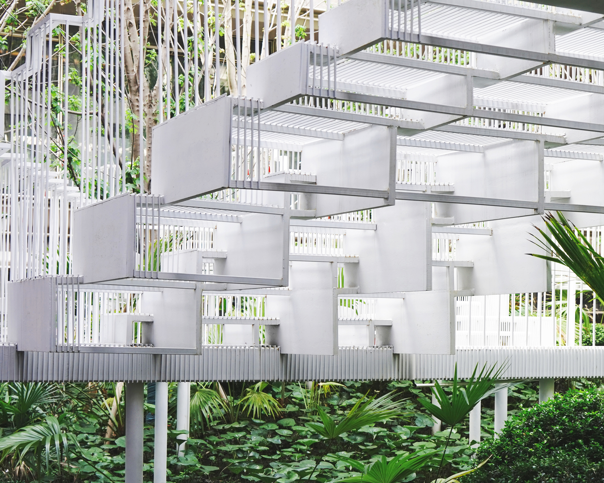 Stacked white box-like steel frames cantilevering above a groundcover bed of round leaves and palm fronds on slender columns