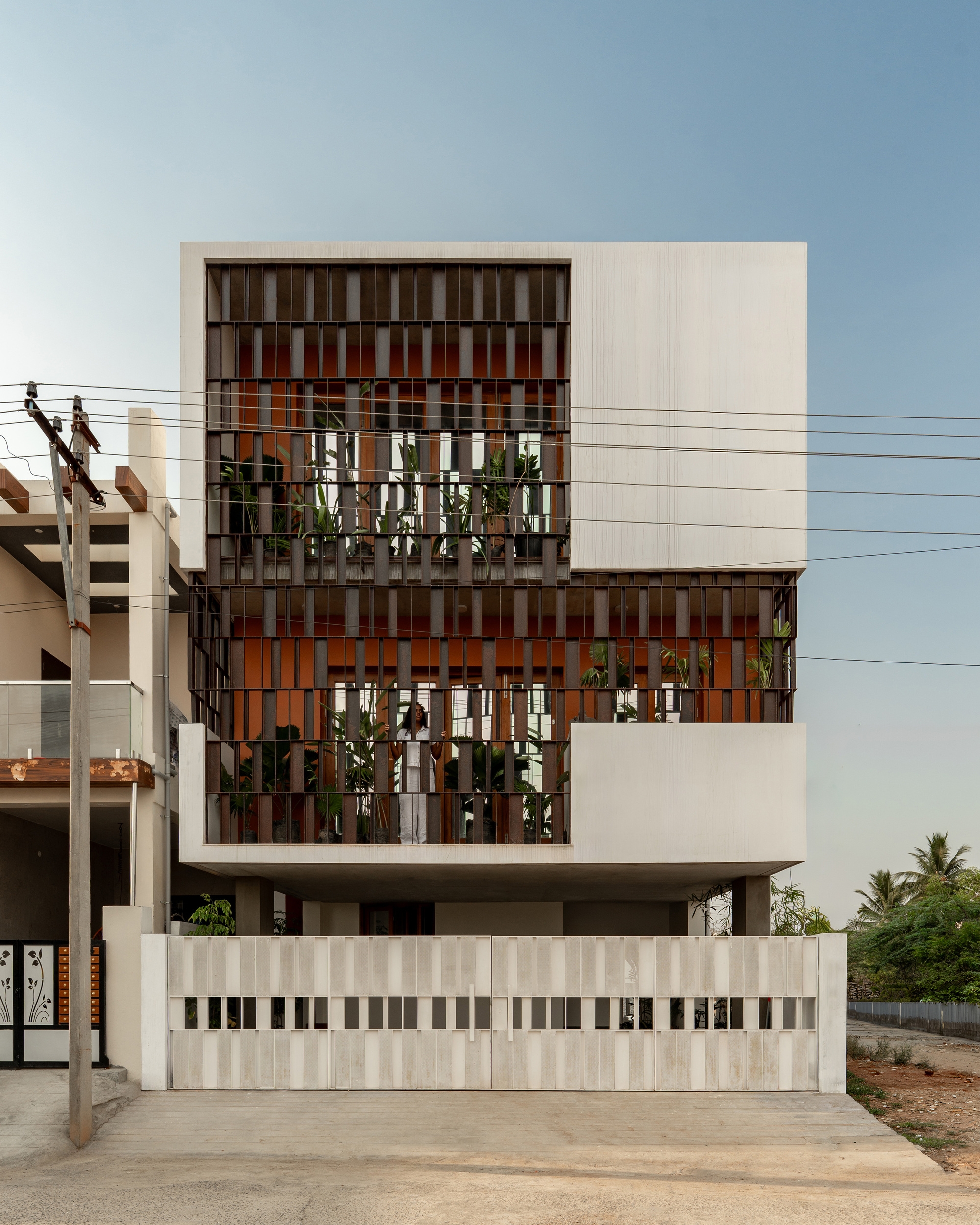 Front elevation of a white concrete house with timber fin screens over planted terraces, framed by overhead power lines at dusk