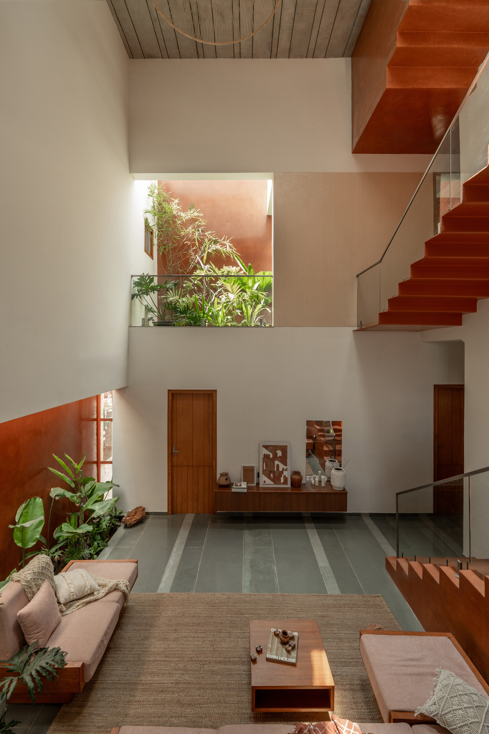 Double-height living room with terracotta-toned stair, indoor plants and a timber door, lit by soft daylight from a skylight