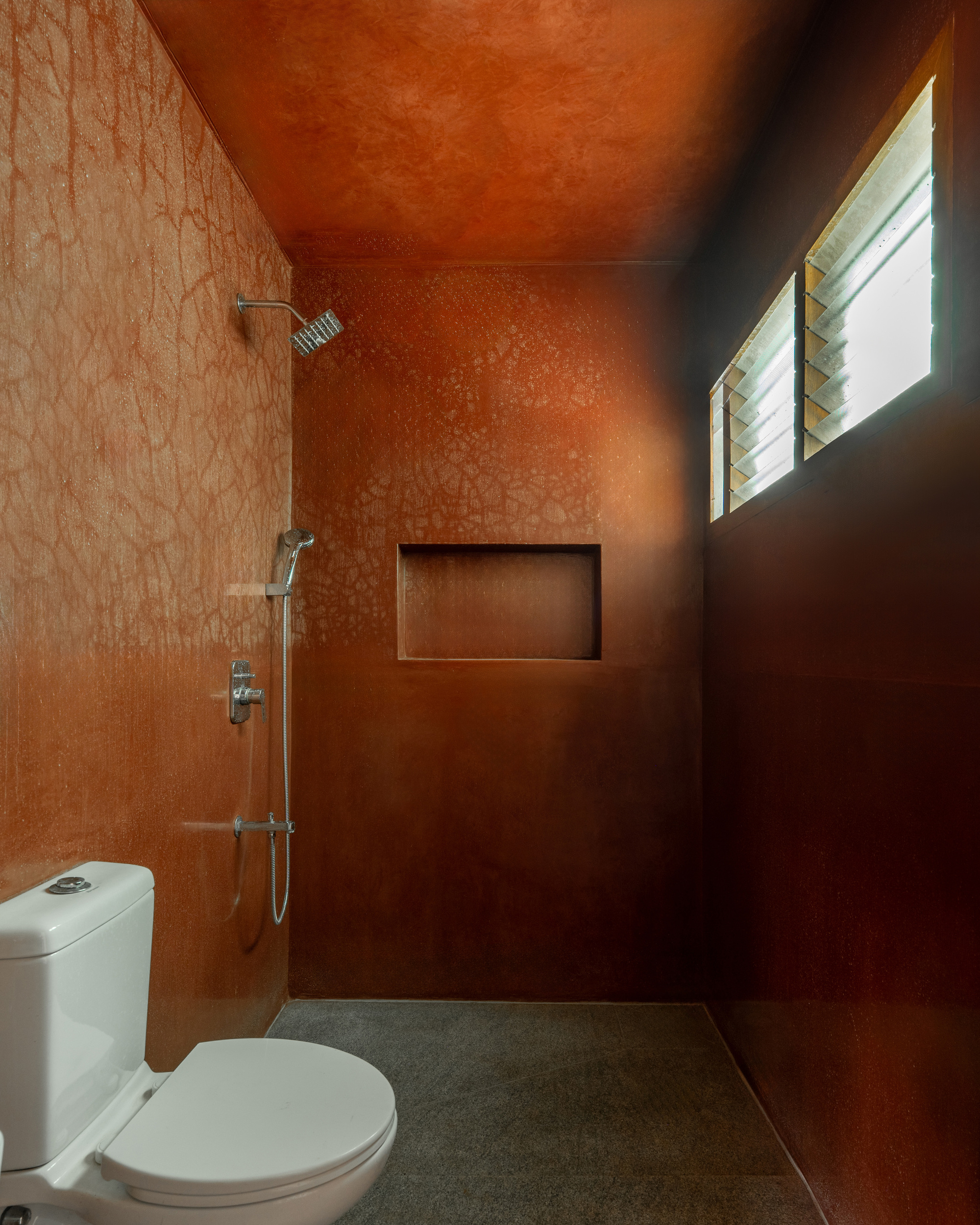 Bathroom lined in rust-coloured textured plaster with a recessed niche and louvred window, daylight glowing through the slats