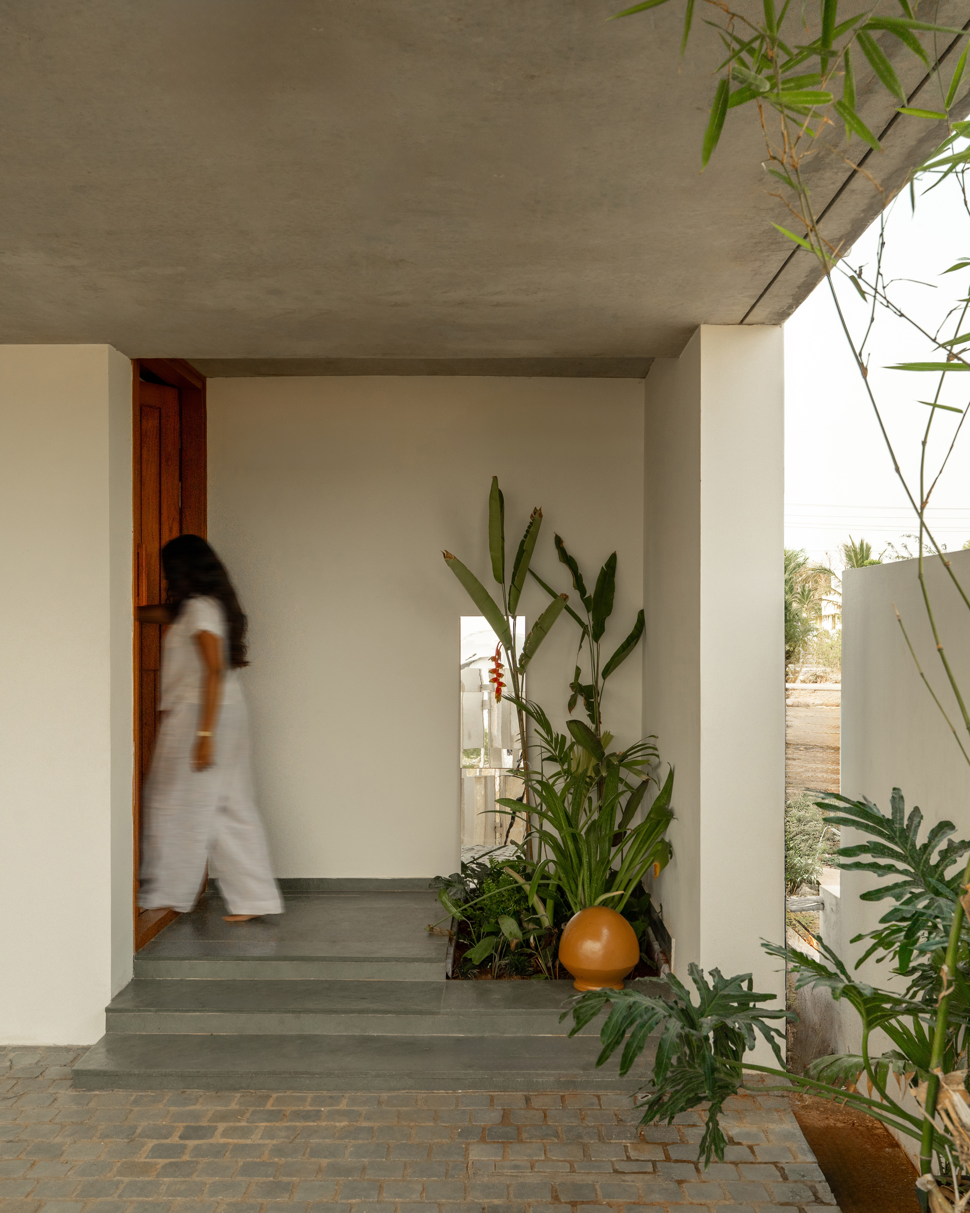 Entry porch with stone steps and a timber door, a blurred woman walking in past a planted bed and orange pot
