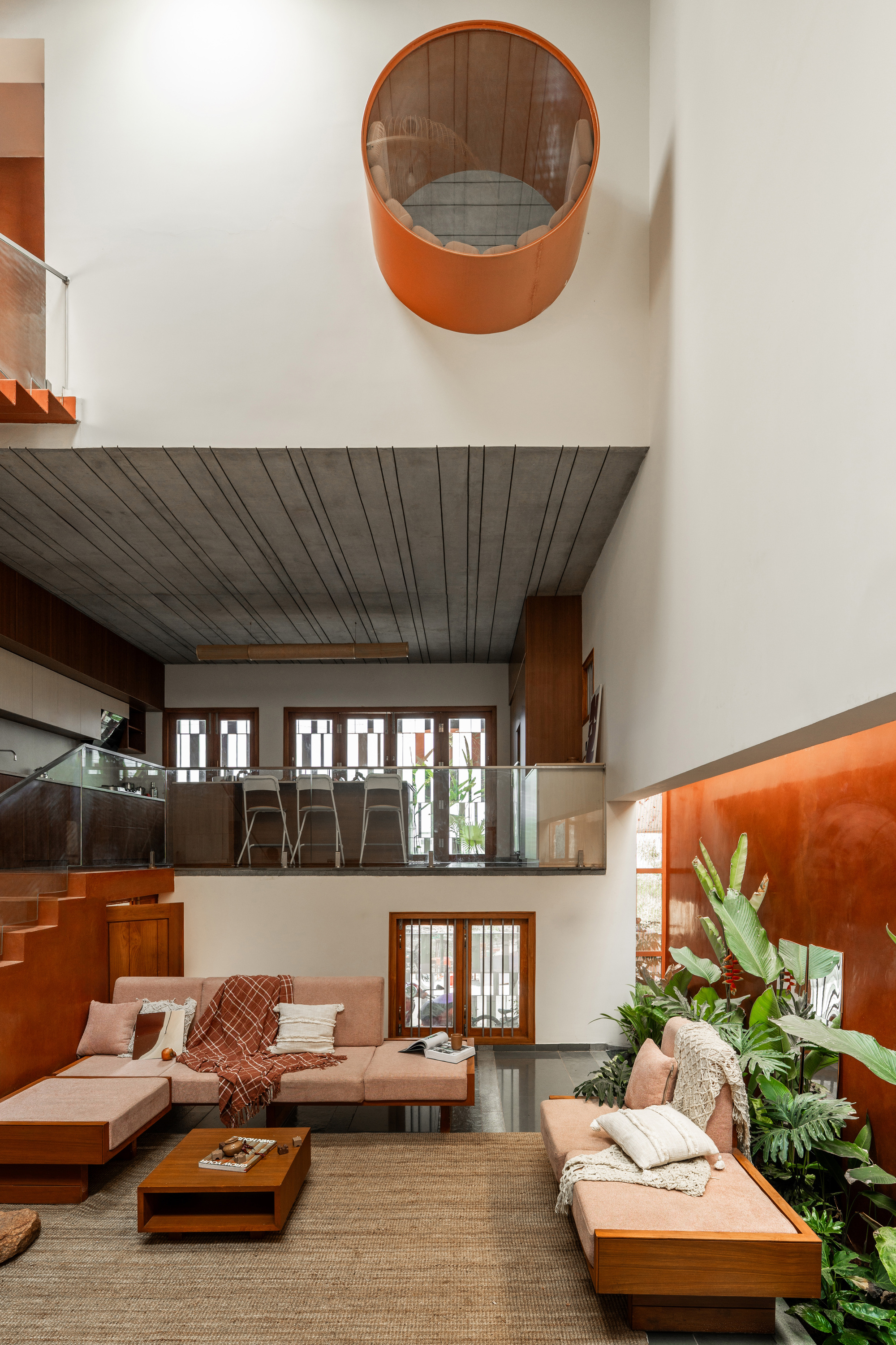 Double-height living room with pink sofas, a round orange window pod overhead and a planted niche against a terracotta wall