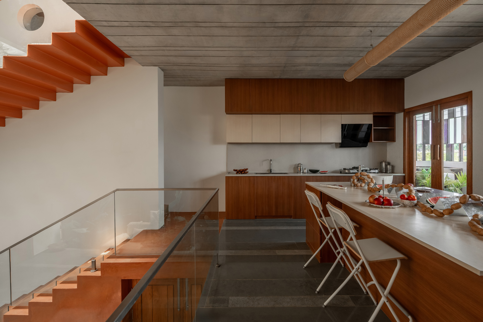 Kitchen with timber cabinetry and a long island with folding stools, beside an orange cantilevered stair and glass balustrade
