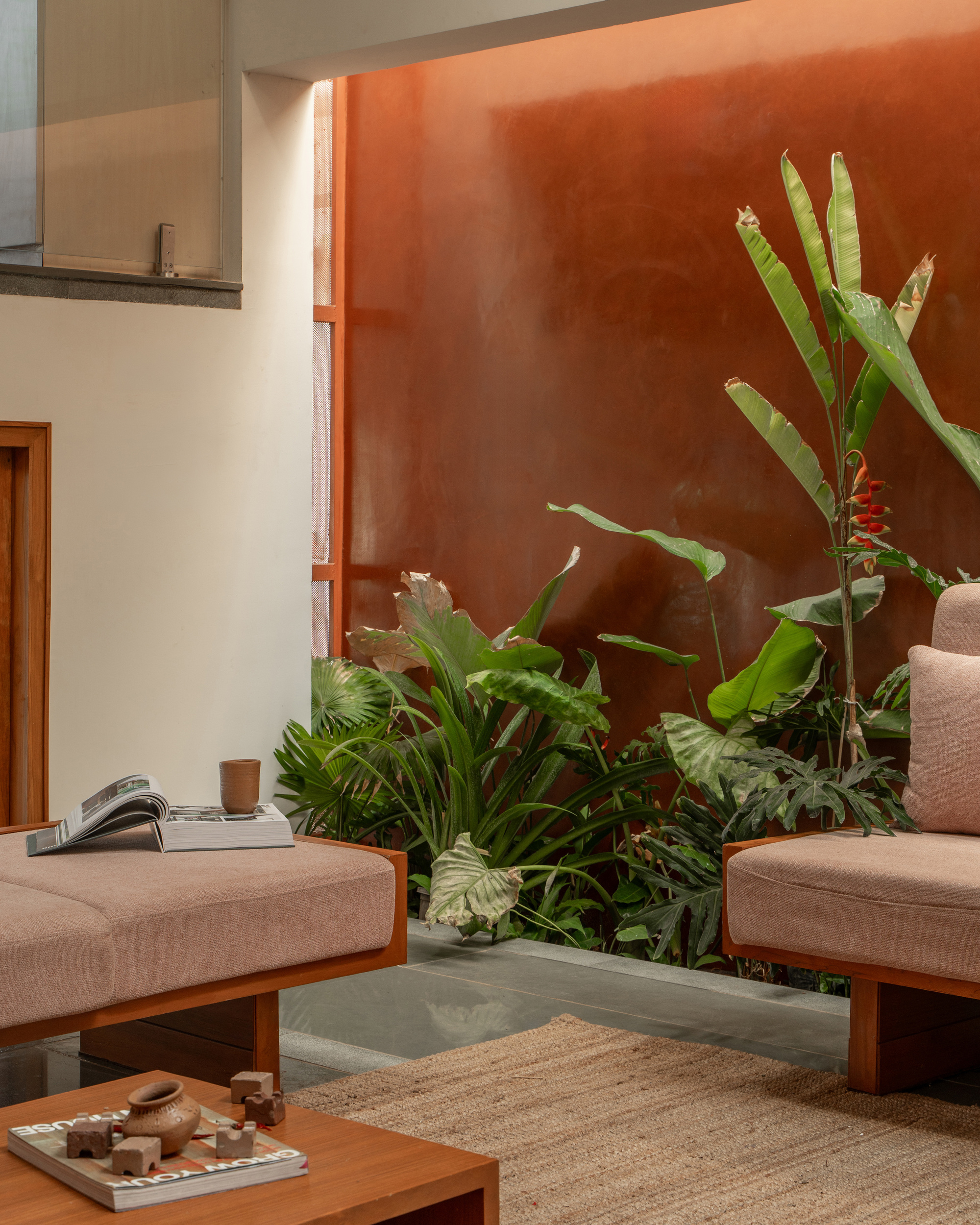Close view of banana plants and ferns against a terracotta plaster wall, with low pink sofas and a jute rug in soft light