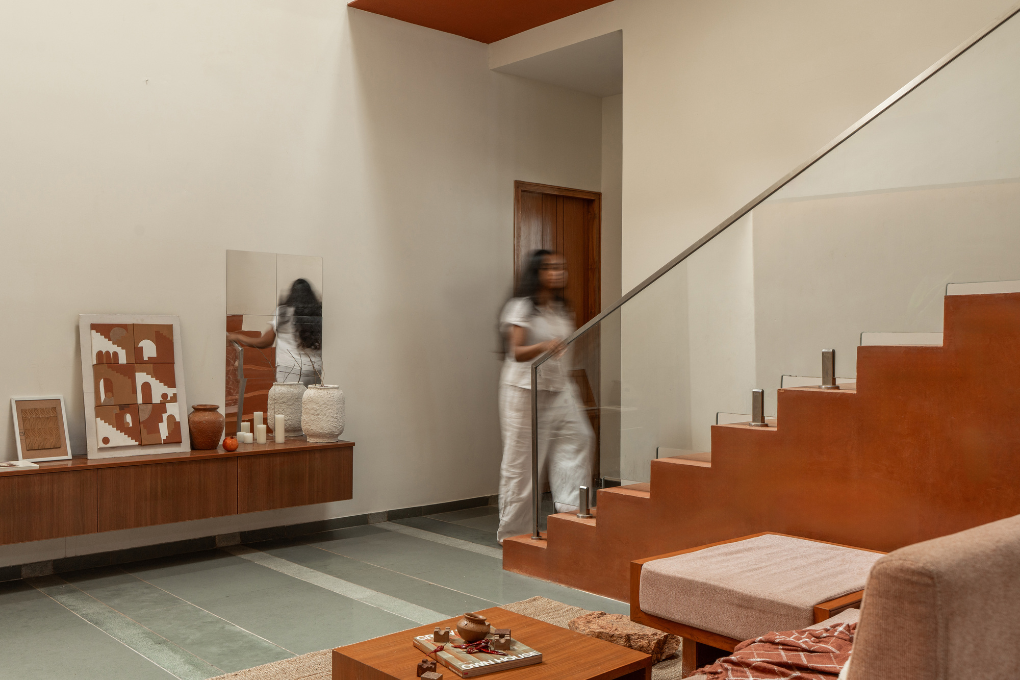Terracotta-toned staircase with glass balustrade beside a wall-hung timber shelf, a blurred figure in white walking past a mirror