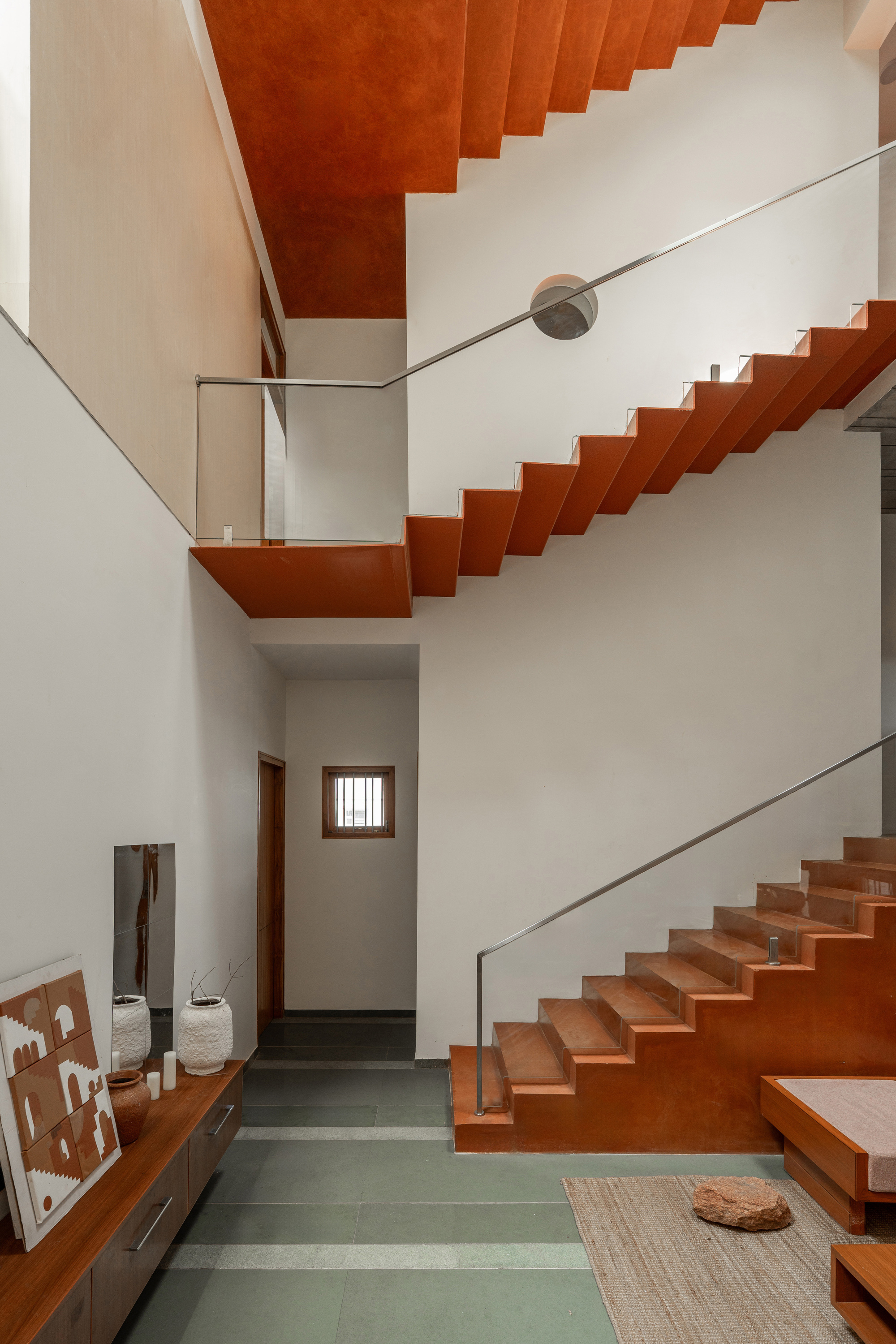 Double-height stair hall with zigzag folded terracotta flights and a steel handrail above a green stone floor and jute rug