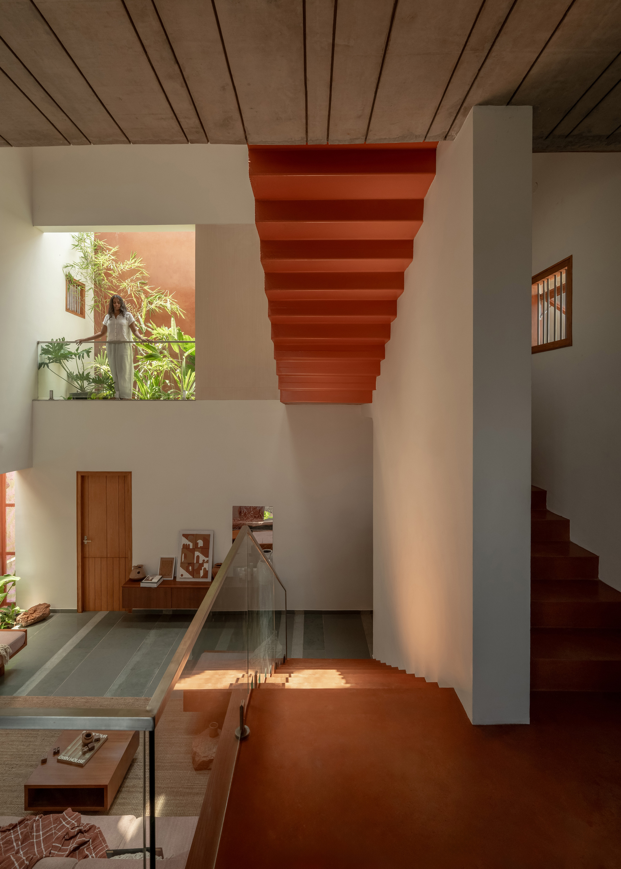 Terracotta stair soffit hanging over a split-level living space, with a figure on an upper landing among tropical plants in daylight