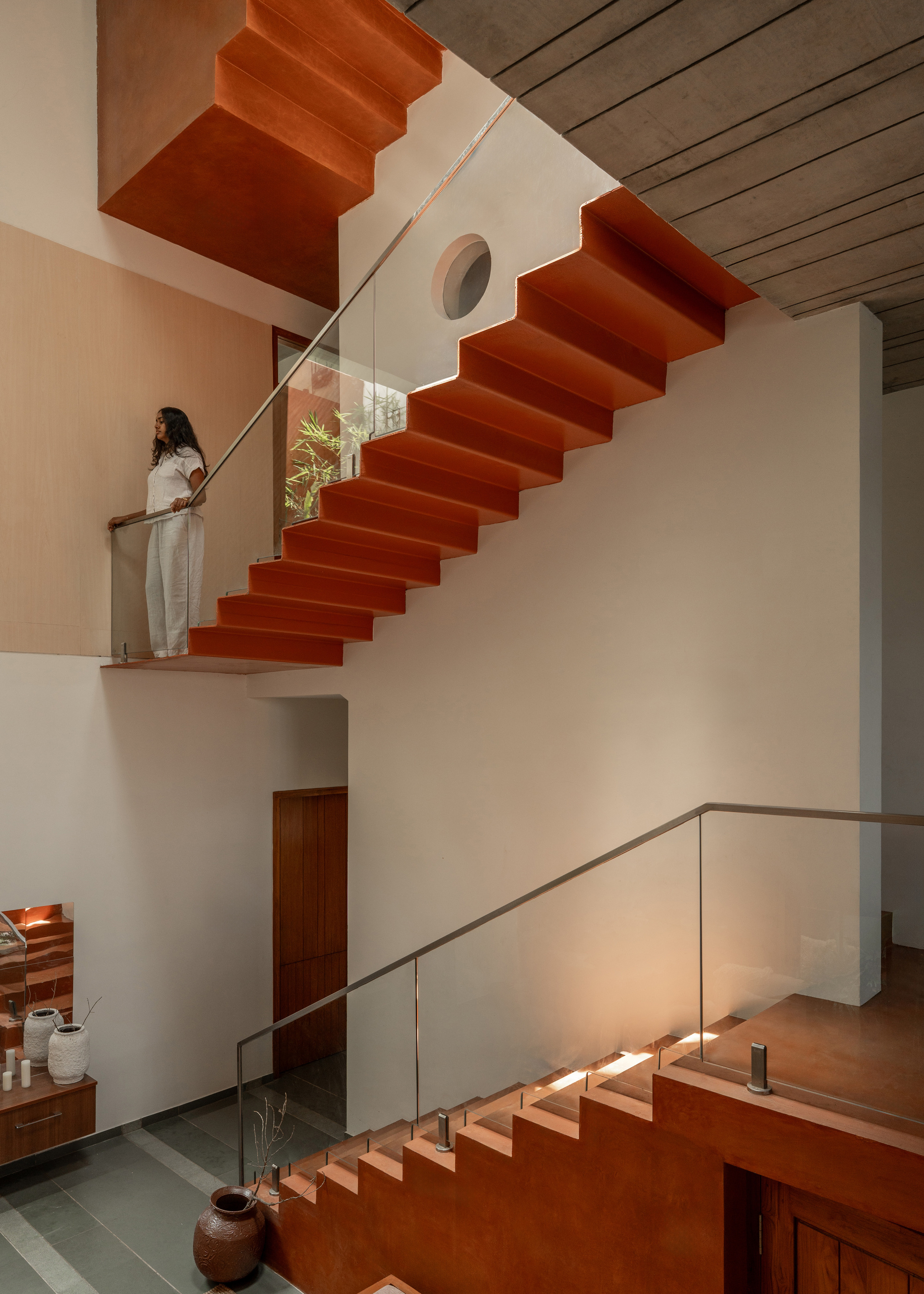 Sawtooth terracotta stair flight crossing a white wall with a round window, a woman standing at the glass-railed landing