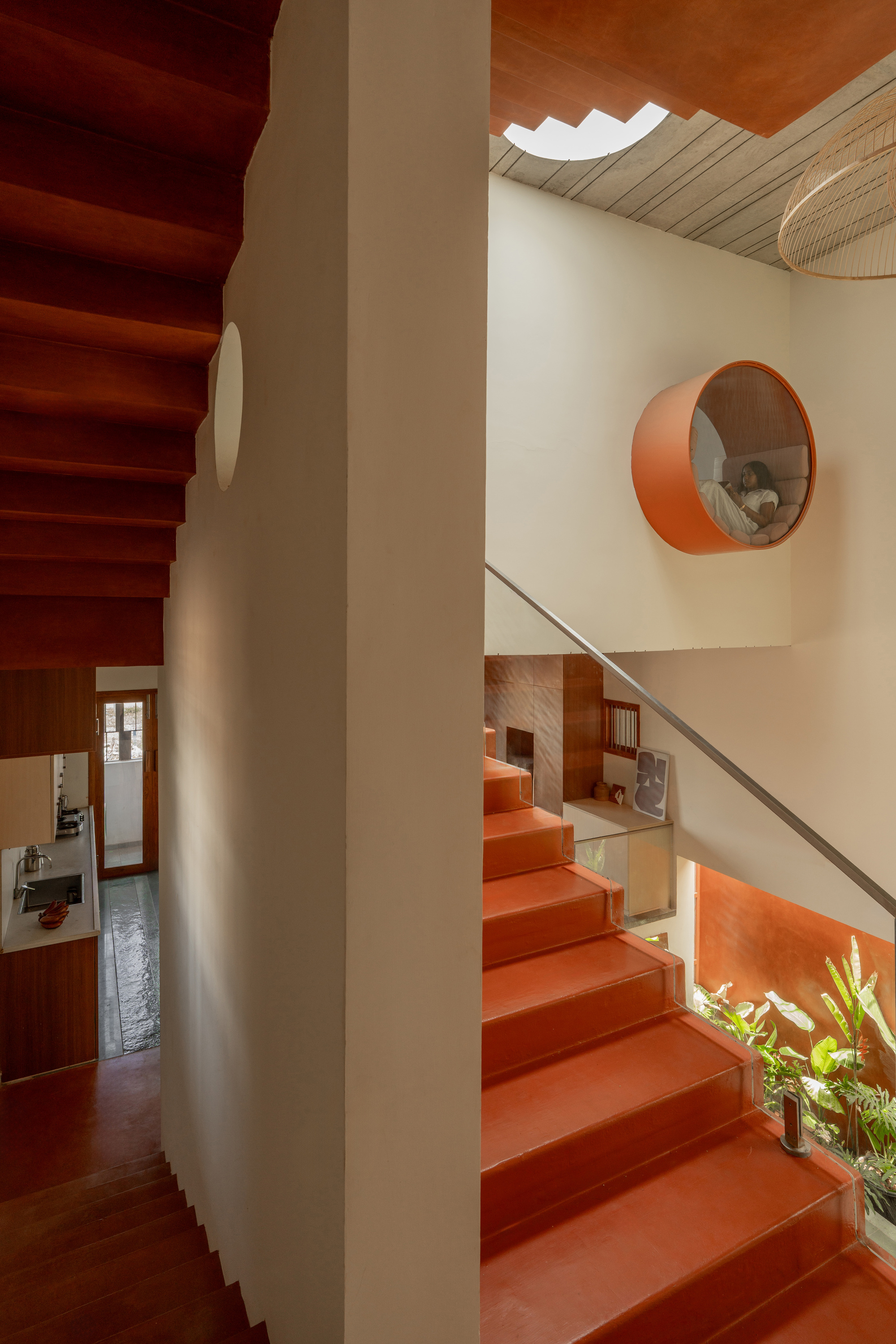 Circular orange reading nook set high in the wall above a terracotta stair, a person seated inside beside planted greenery