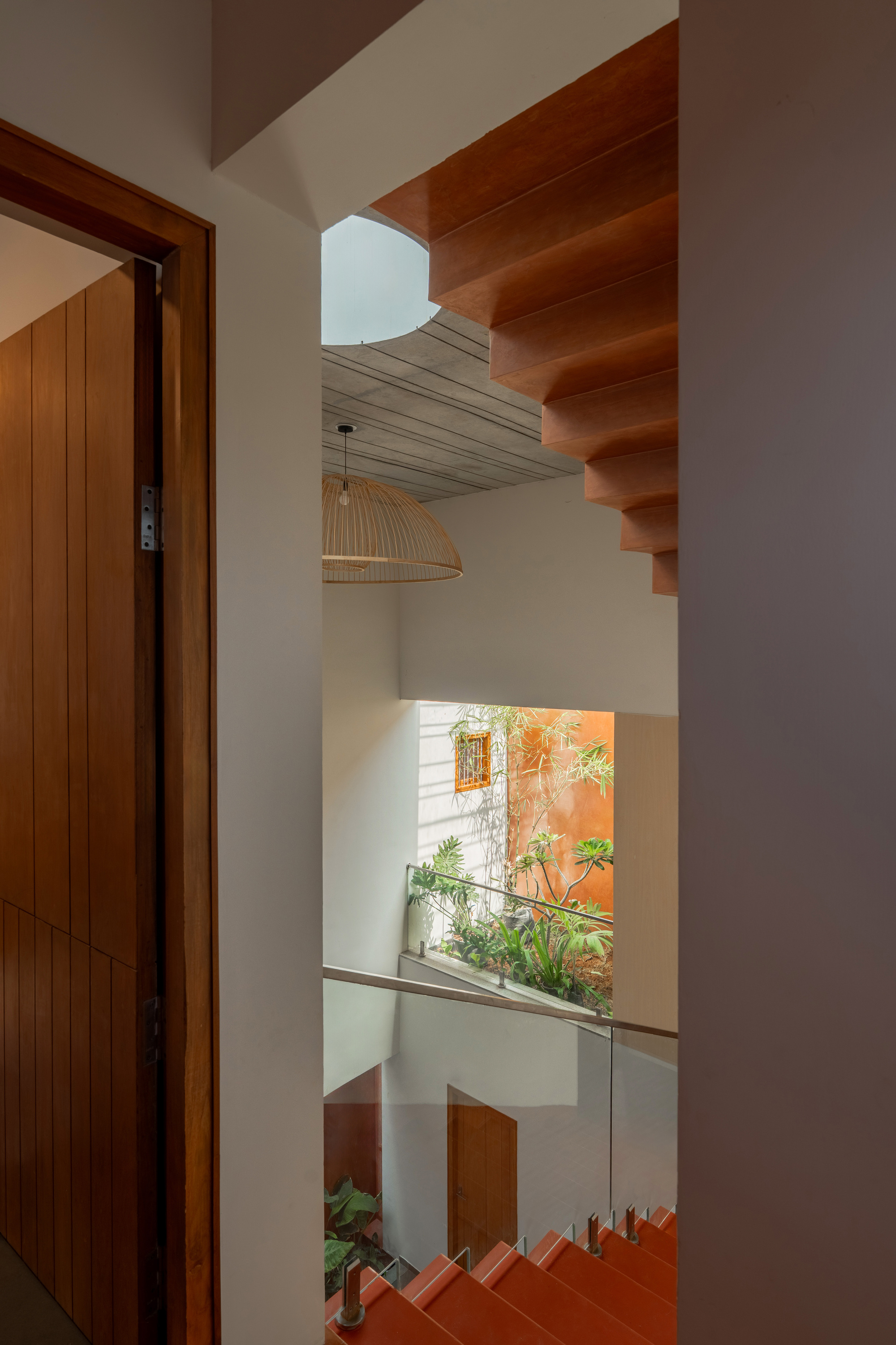 View through a timber door down a stairwell to a planted light well, a woven pendant lamp hanging below a round skylight