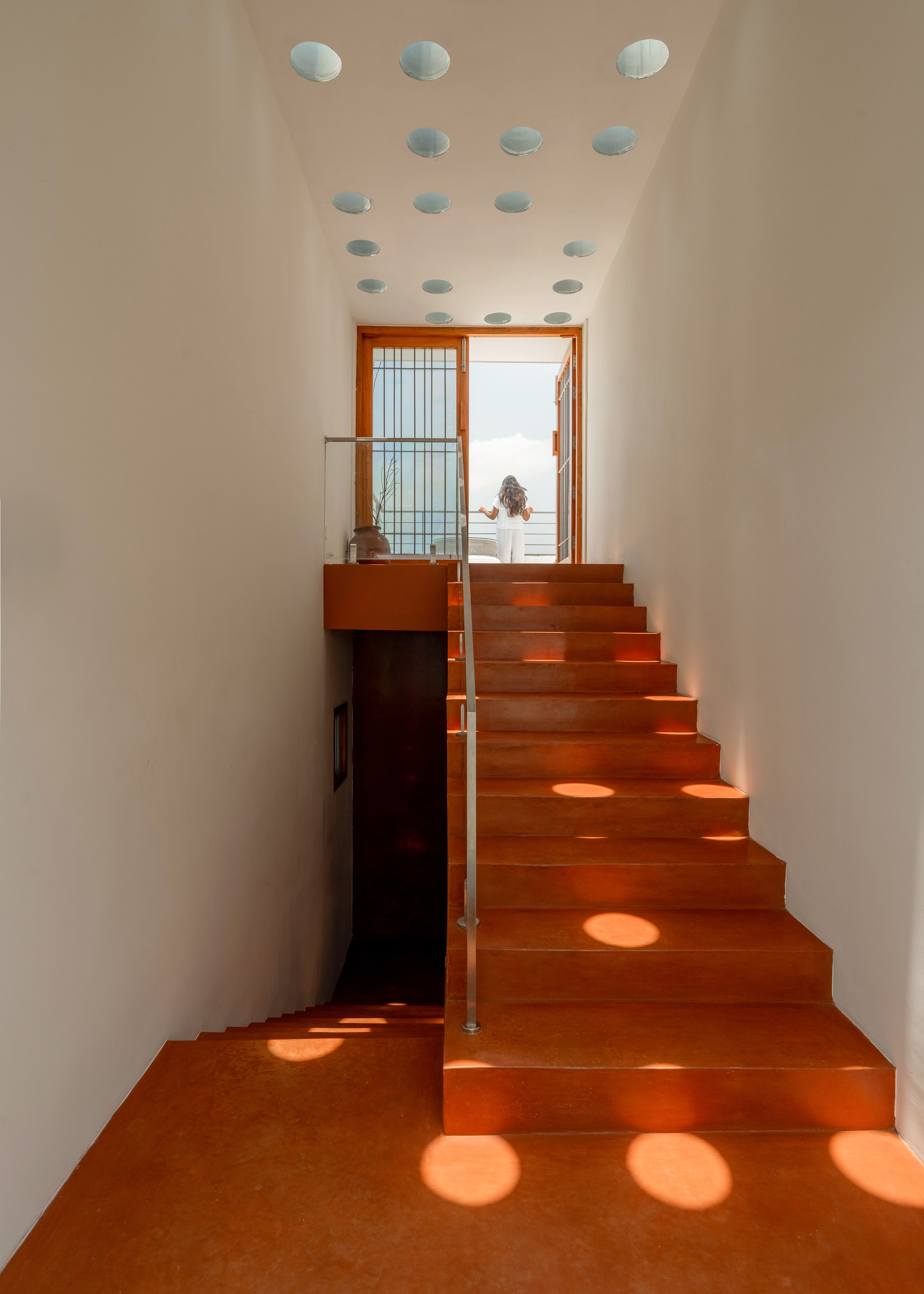 Terracotta-toned stair rising toward a balcony door, with round skylight openings casting discs of sunlight across the steps and floor