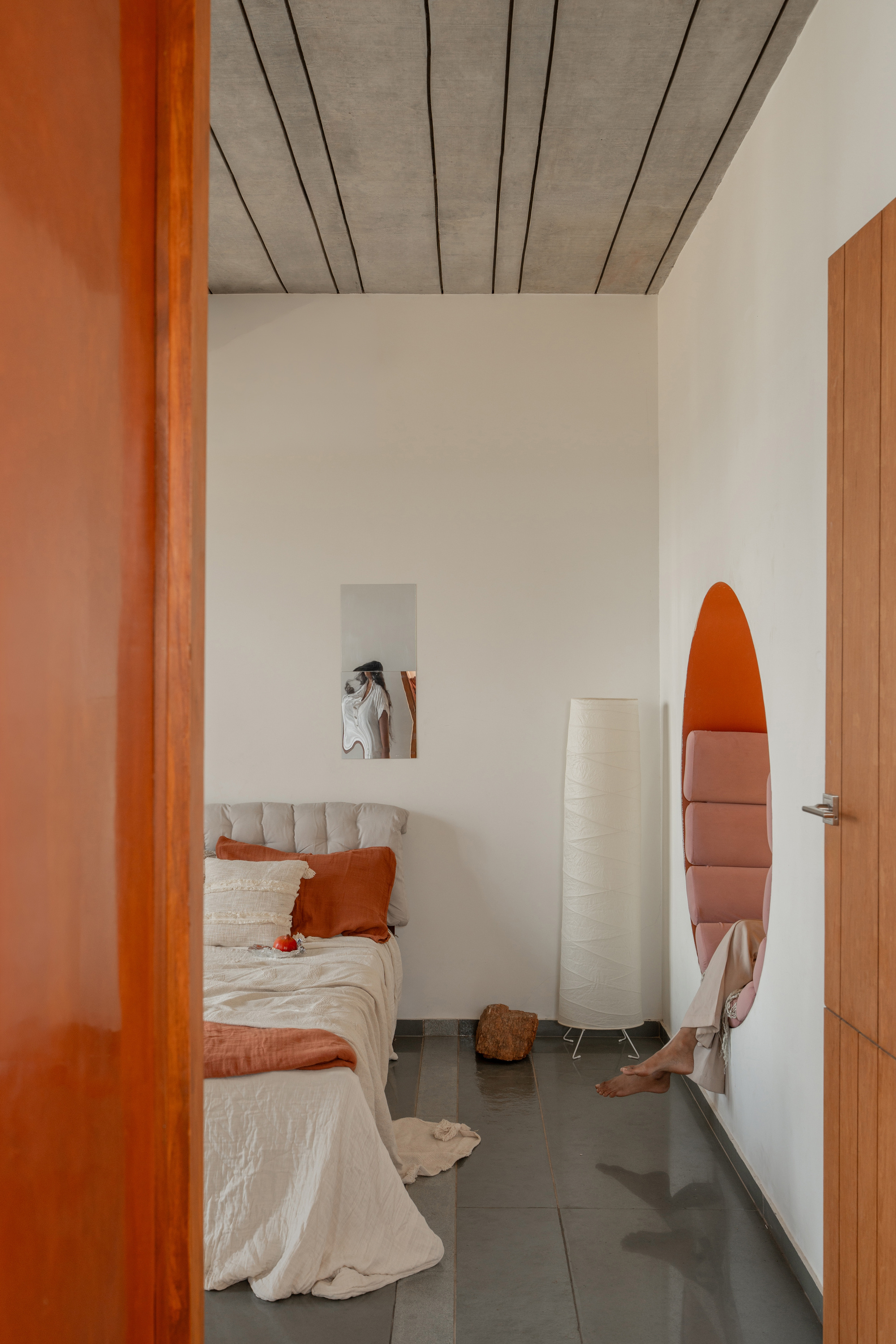 Bedroom seen through an open timber door, with a round orange wall niche holding a person's bare feet and a concrete plank ceiling