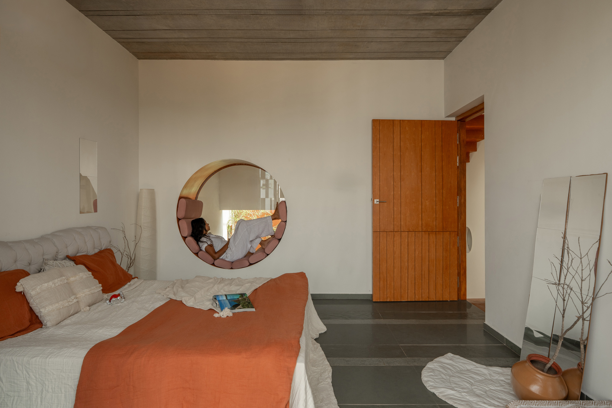Bedroom with a circular cushioned window seat in the white wall, a person reclining inside, and a vertical plank timber door beside it