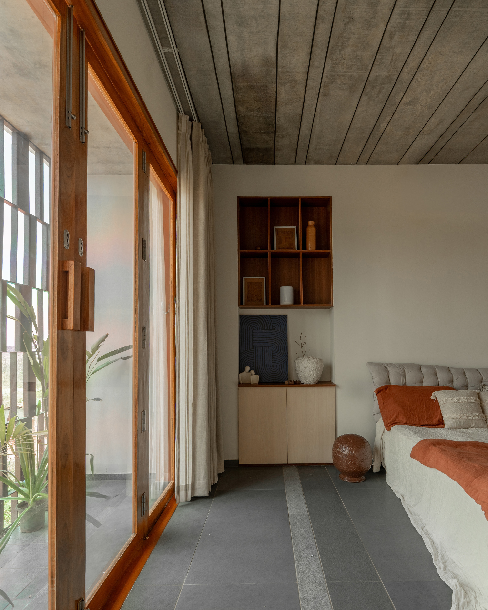Sliding timber-framed glass doors open to a planted balcony beside a bed, with built-in shelves and a stone-inlaid grey floor