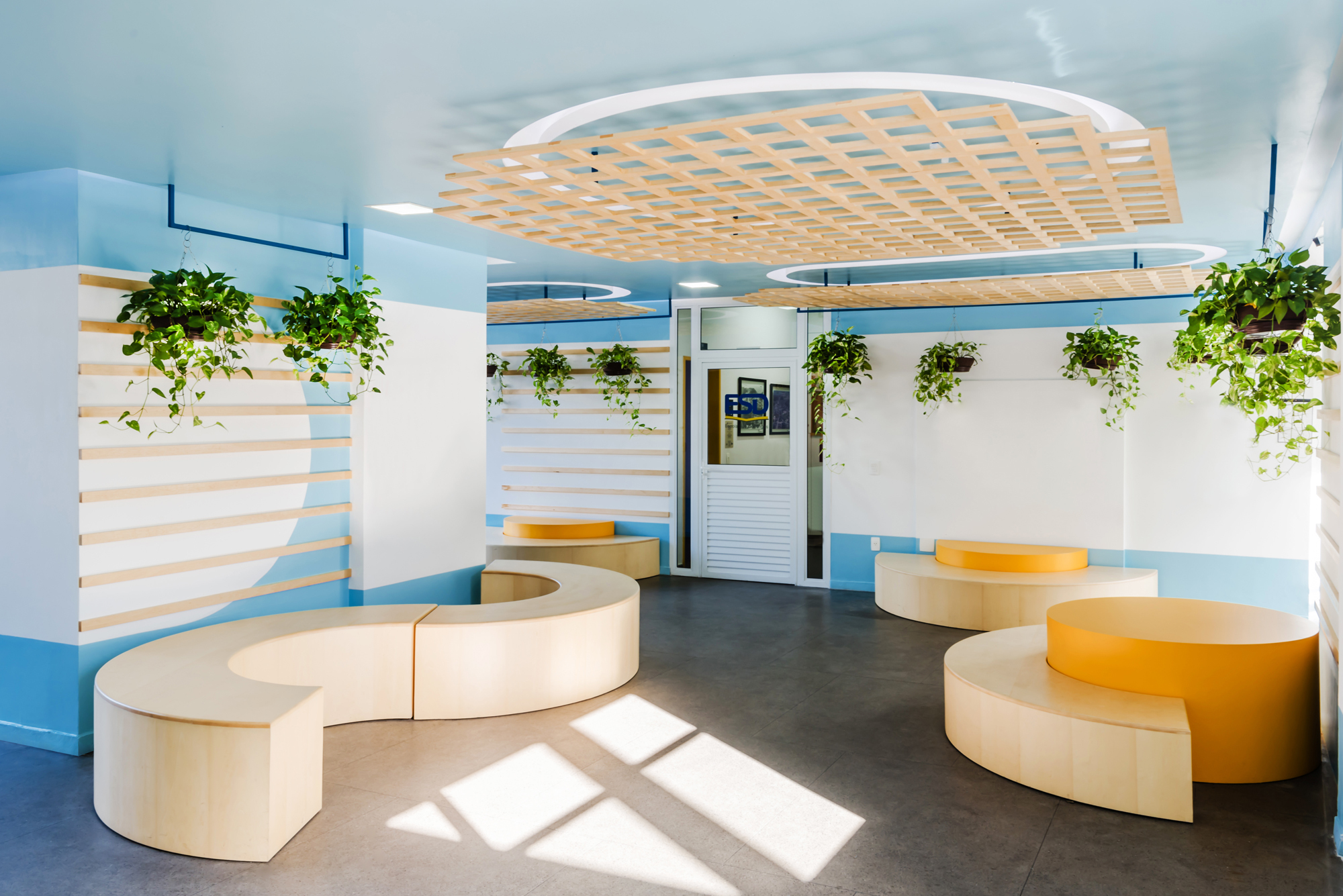 Waiting area with curved timber benches, hanging pothos plants and a circular lattice ceiling panel, with sunlight patches falling across the grey floor