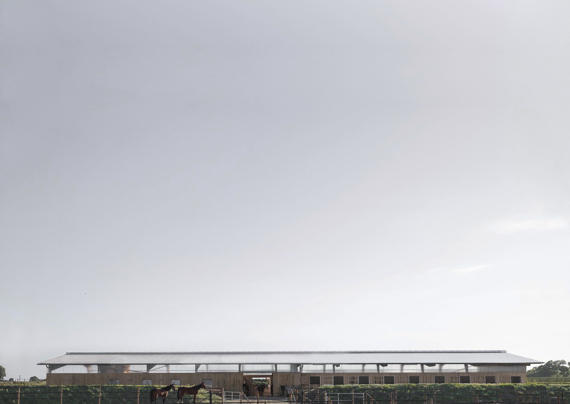 Distant long view of the low barn with pale roof across a field fence, two horses grazing under an overcast sky