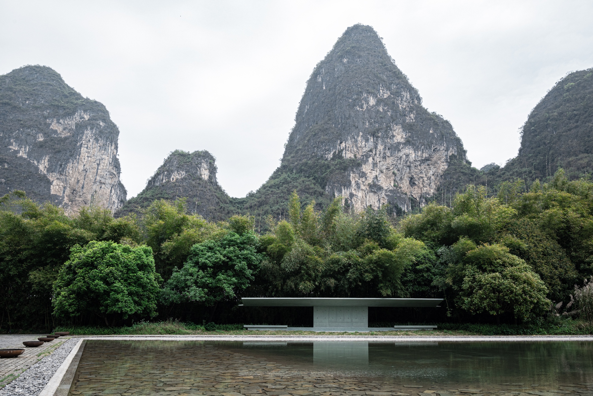 Low concrete pavilion across a rectangular pond, with limestone peaks rising behind the bamboo under an overcast sky