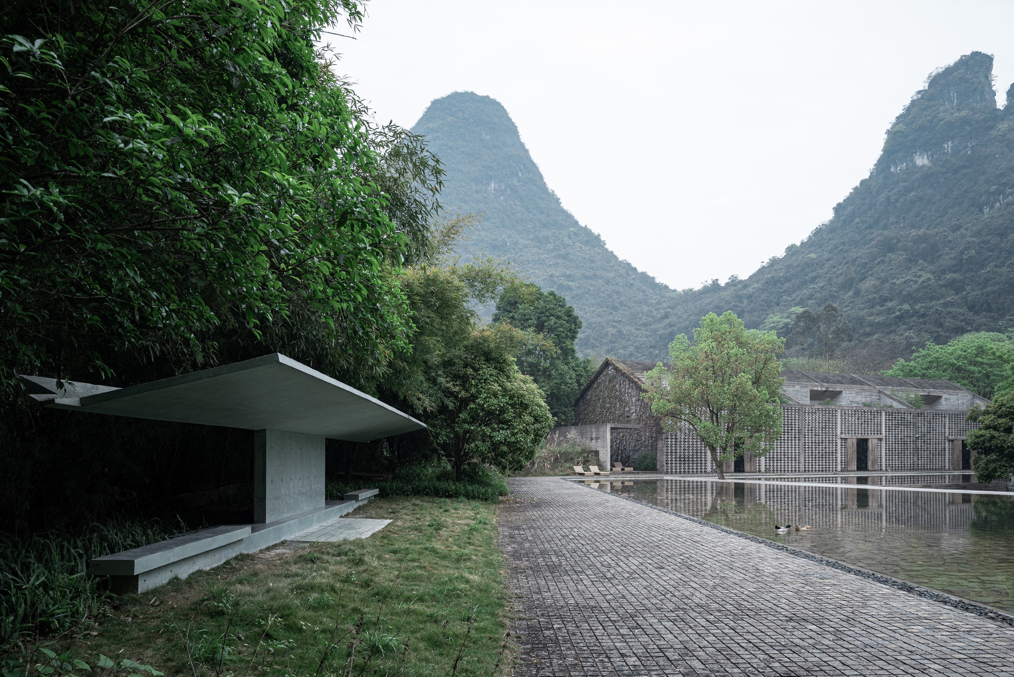 Cantilevered concrete shelter beside a paved path leading to a stone-clad building and pond beneath misty karst hills