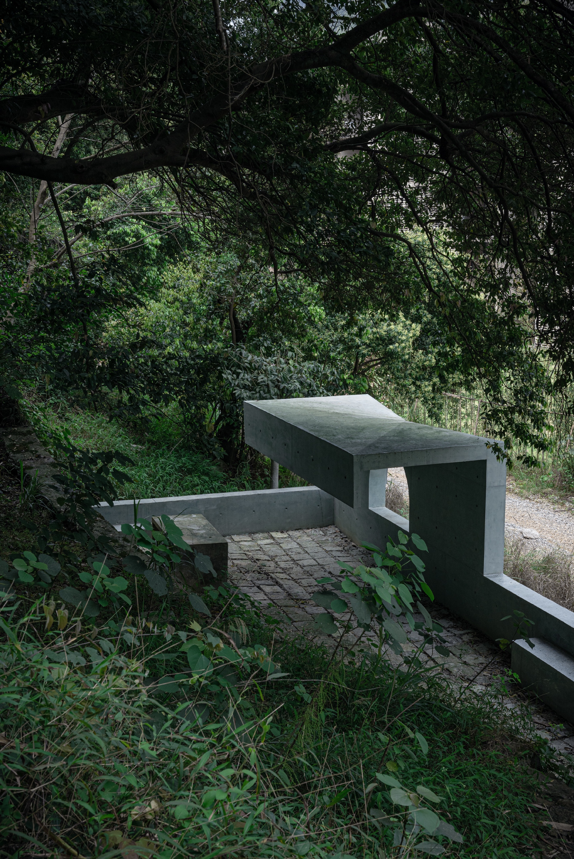 Concrete bench and cantilevered slab on a brick-paved terrace, half hidden under overhanging branches and ferns