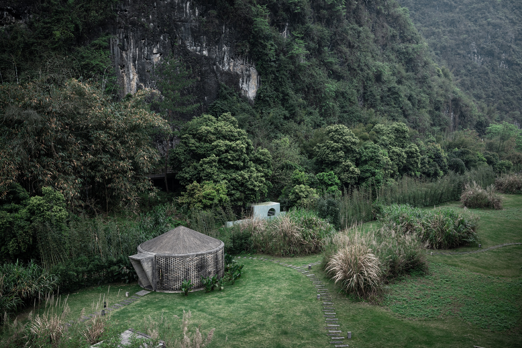 Aerial view of a round thatched hut with lattice walls on a lawn below a rocky green hillside