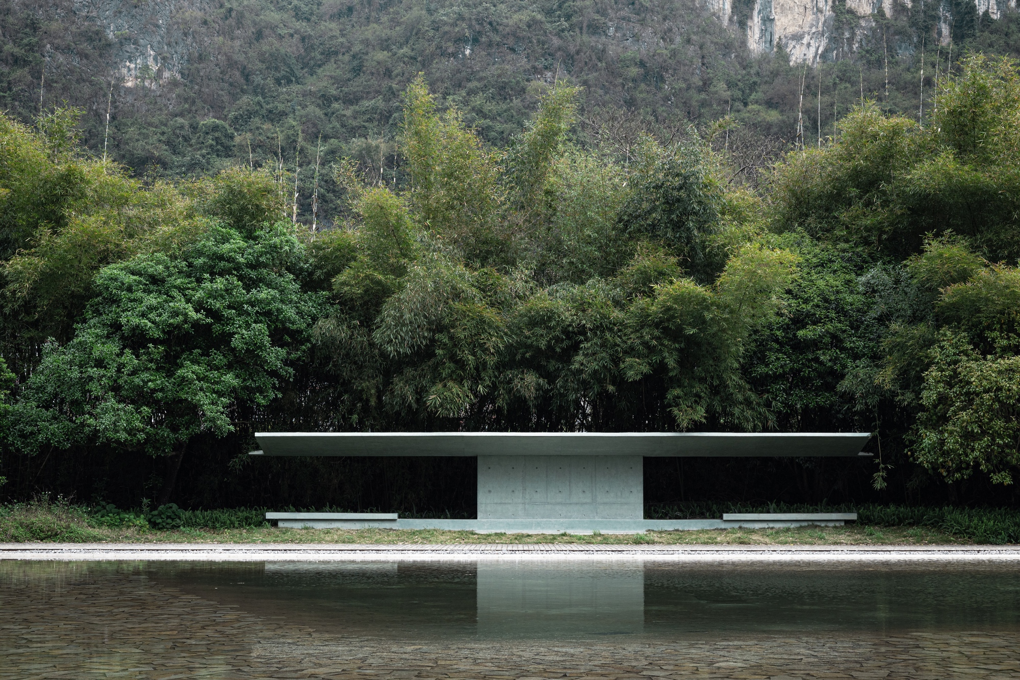 Flat concrete canopy on a single central pier beside a still pond, backed by dense bamboo and a limestone hillside