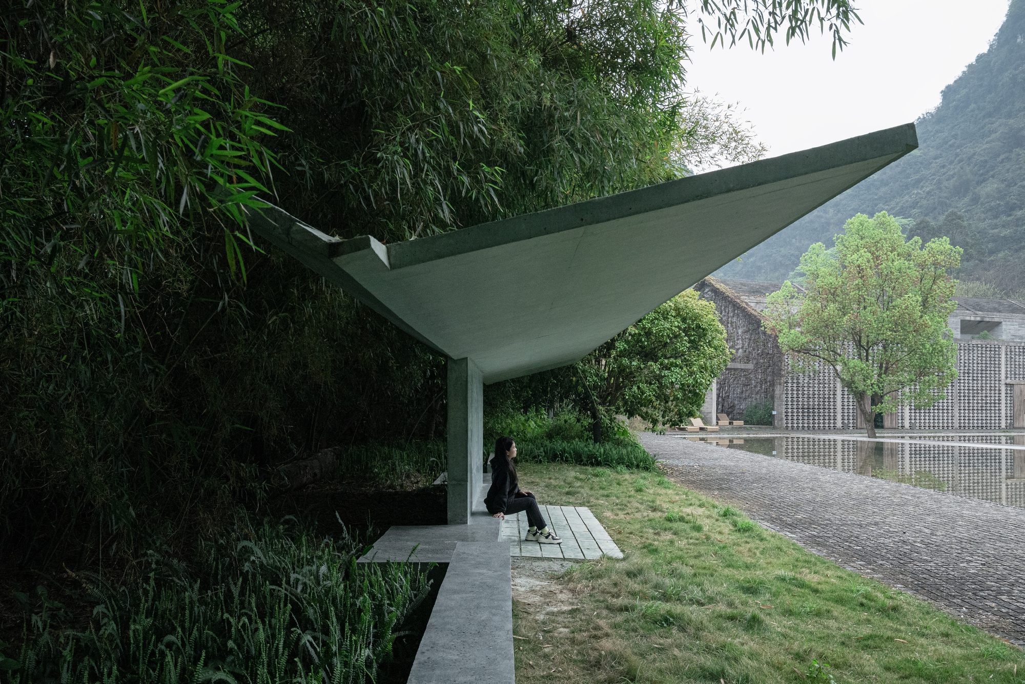 Person seated under the tilted concrete canopy, with a paved lakeside path and a perforated brick building across the water