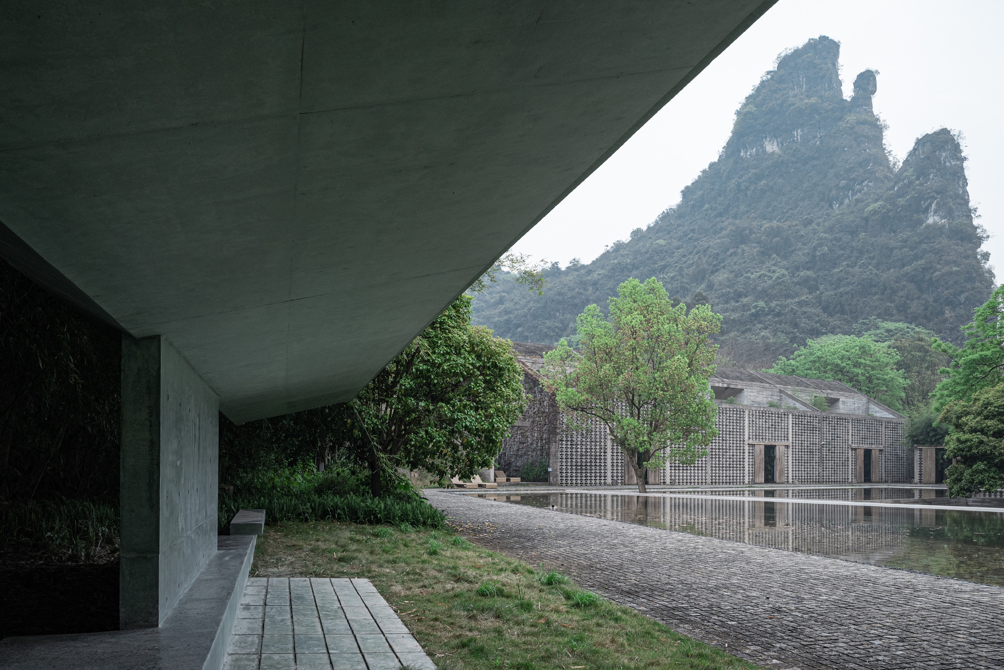 View from beneath the concrete canopy across a reflecting pond to a lattice-walled building and a jagged karst peak in mist