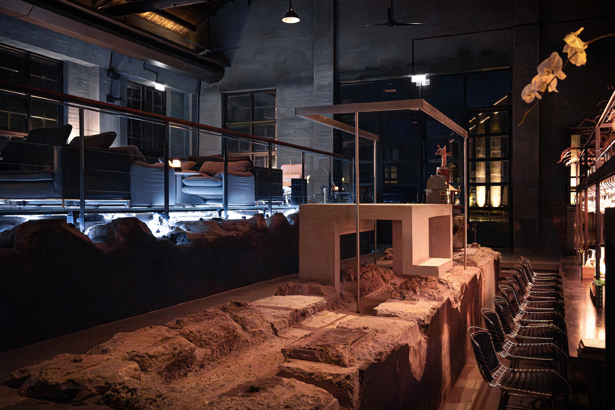 Bar counter set on a rough excavated rock bed, with glass balustrade, lounge seating and wire stools in warm low light