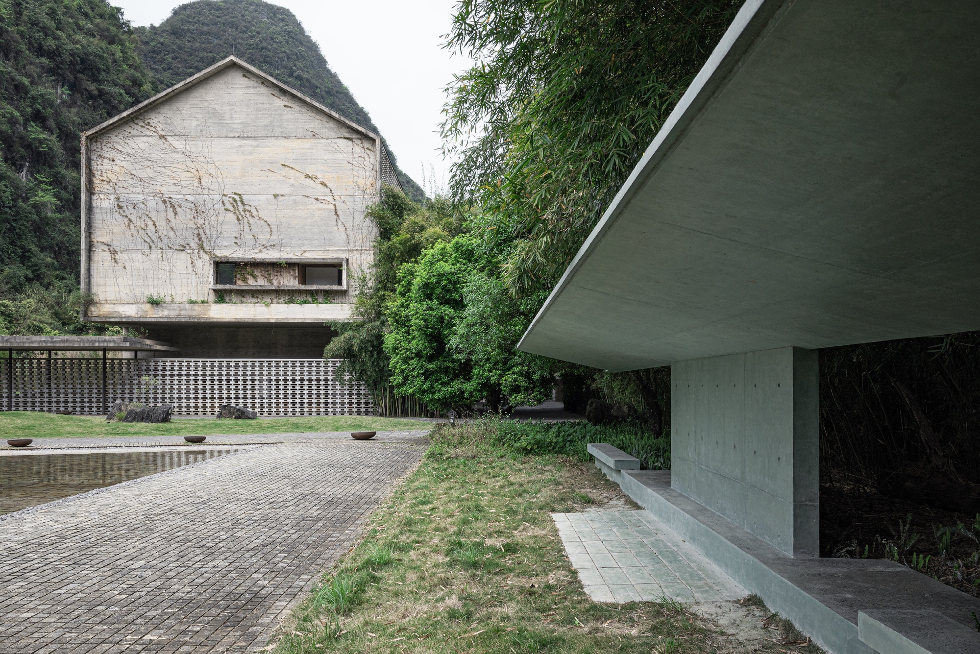 Cantilevered concrete shelter beside a brick-paved court, with a vine-streaked gabled concrete building and limestone hill beyond