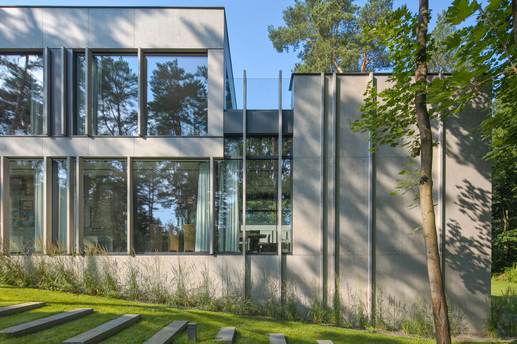 Close view of grey panel facade with vertical fins and tall windows reflecting pines, beside stepping stones in the grass
