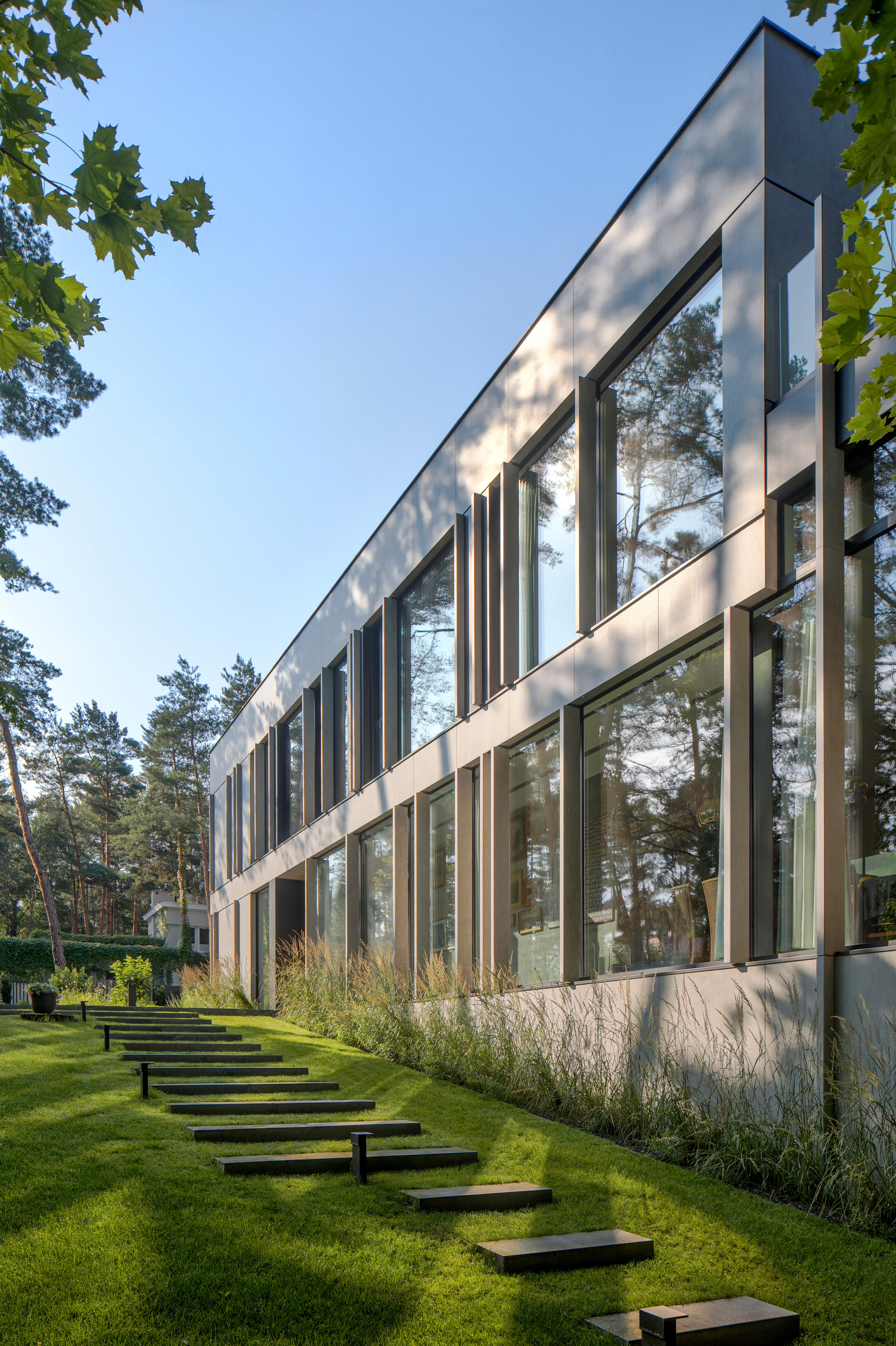 Long side facade with slim vertical mullions and reflective glazing above a line of stepping stones and ornamental grasses in low sun